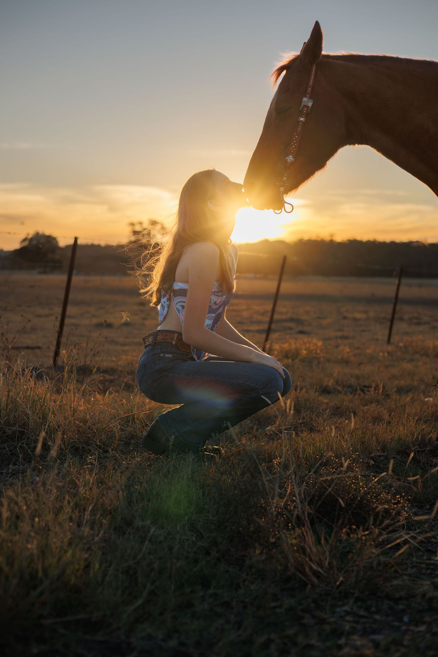 Woman kneeling beside a chestnut horse at golden sunset in a rural field, warm sunlight creating a beautiful lens flare.
