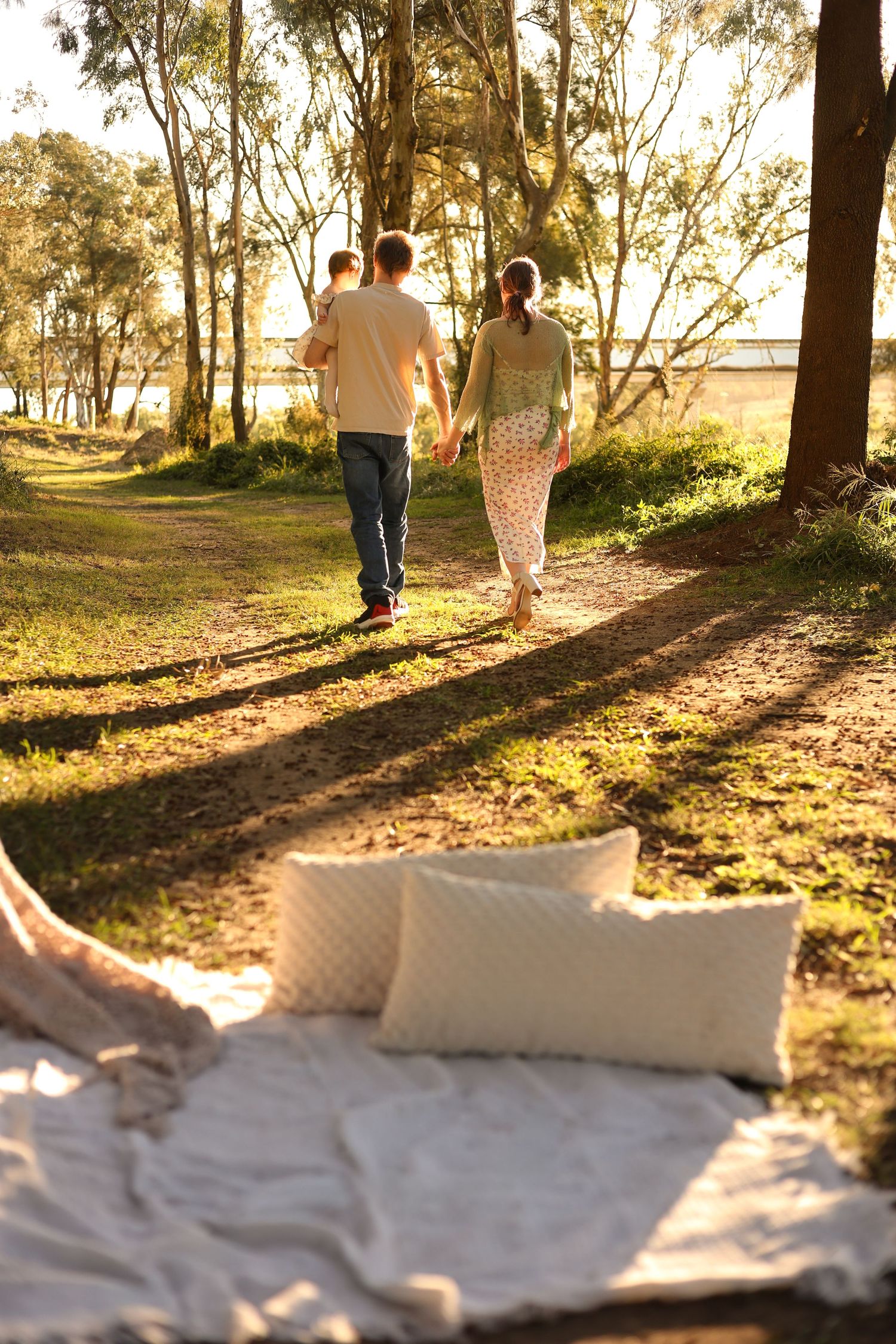 A family walks hand-in-hand through a sunlit park with tall trees, blanket and pillows in foreground.