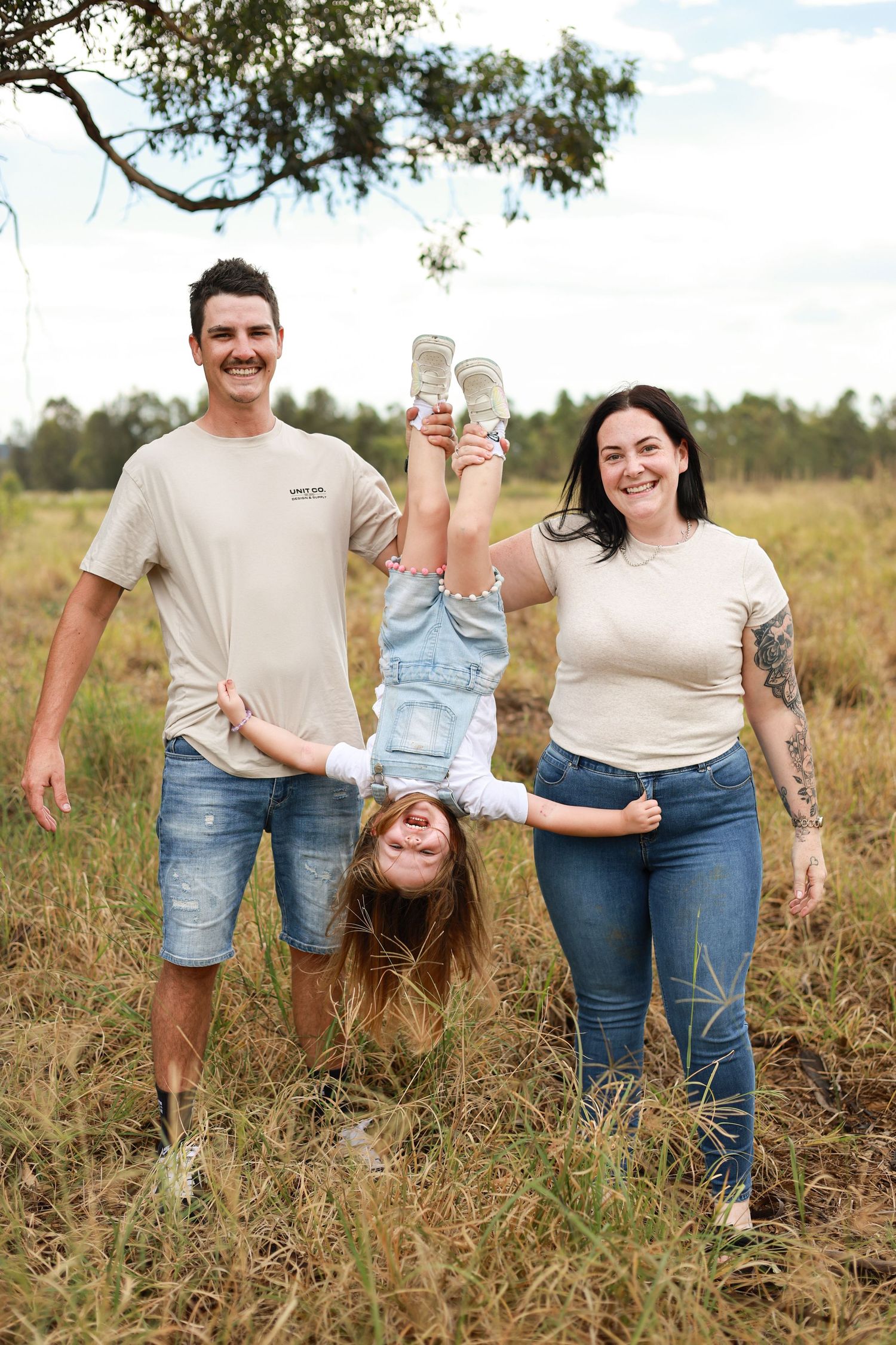 A happy family of three outdoors, parents playfully holding their child upside down in a grassy field.