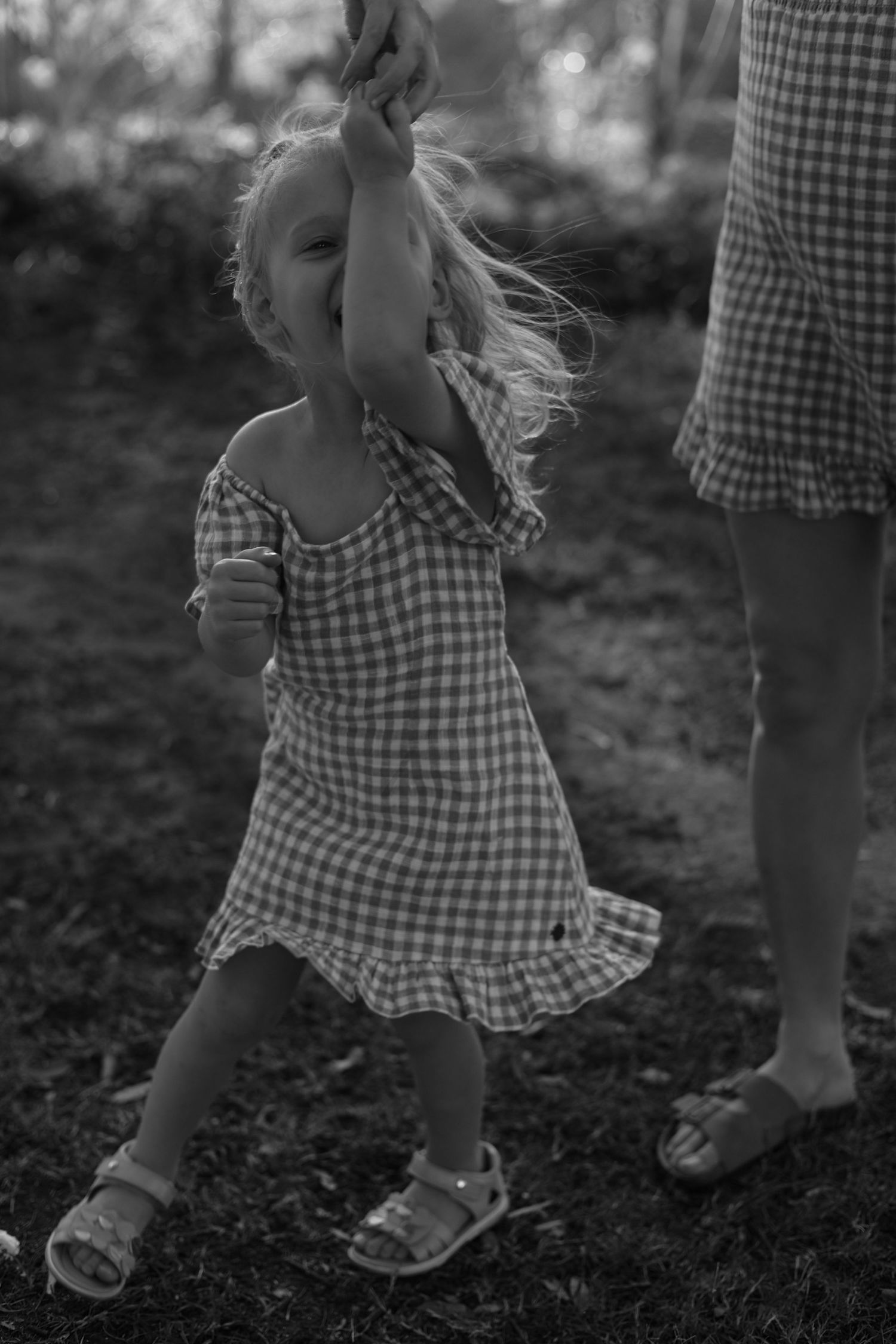 Black and white photo of a toddler girl in gingham dress holding an adult's hand outdoors.