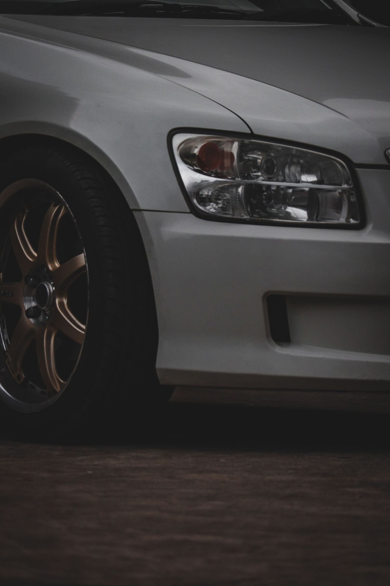 Close-up detail shot of a white car's front end showing headlight and bronze-colored aftermarket wheel.