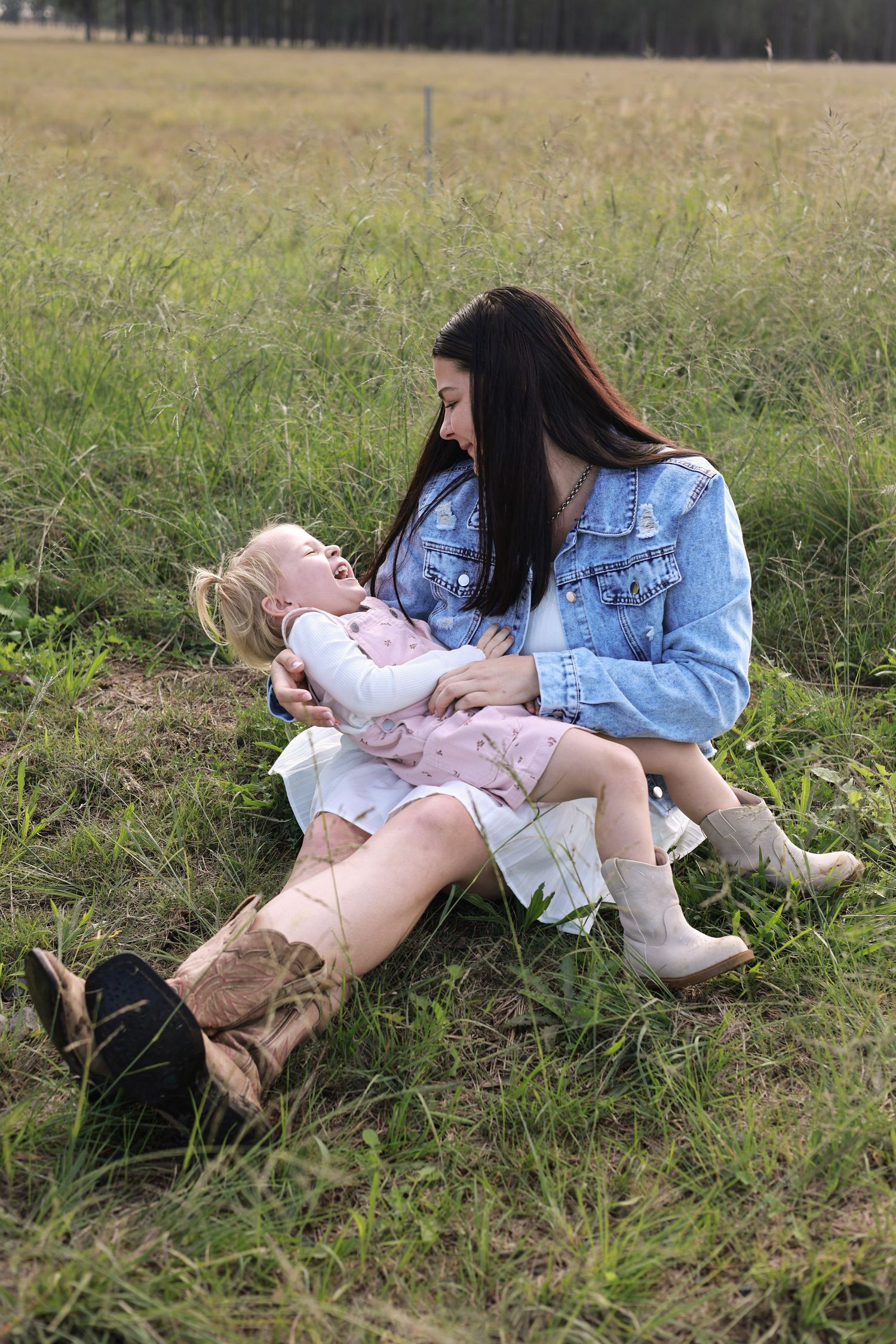 A young mother in a denim jacket sits in a grassy field laughing with her toddler daughter in cowboy boots.