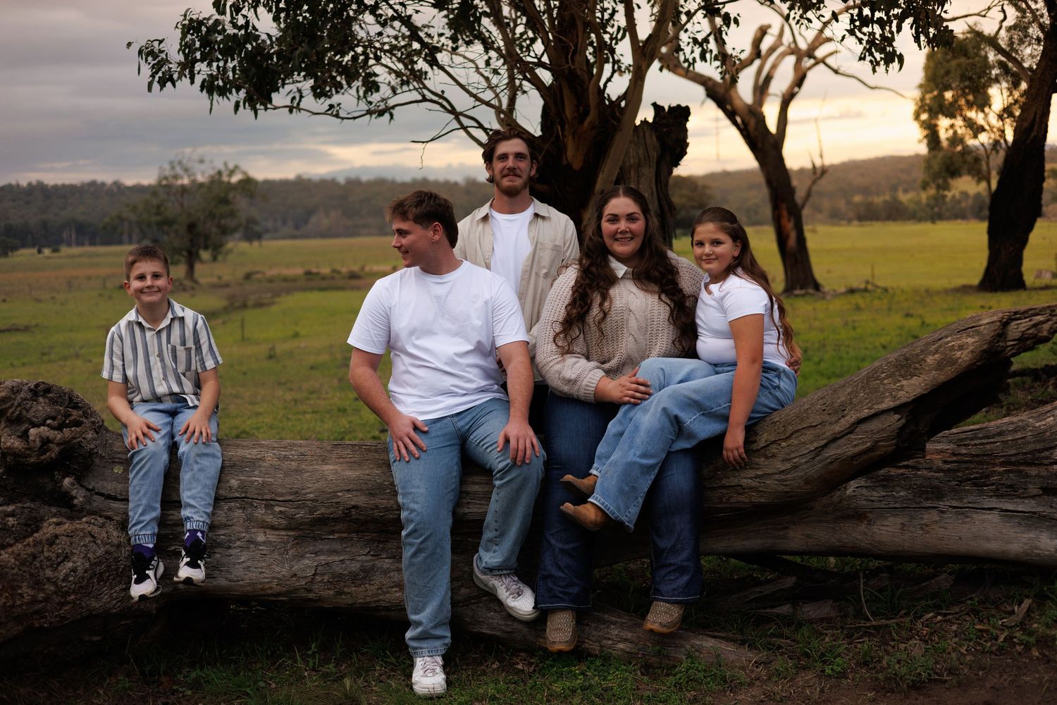 Family of five posing on a fallen log in a lush green Australian countryside setting at dusk.