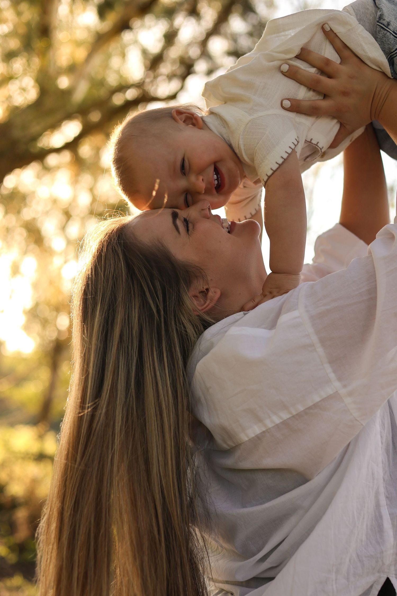 Mother lifting smiling baby in sunlit park, both wearing white, sharing a joyful moment outdoors.