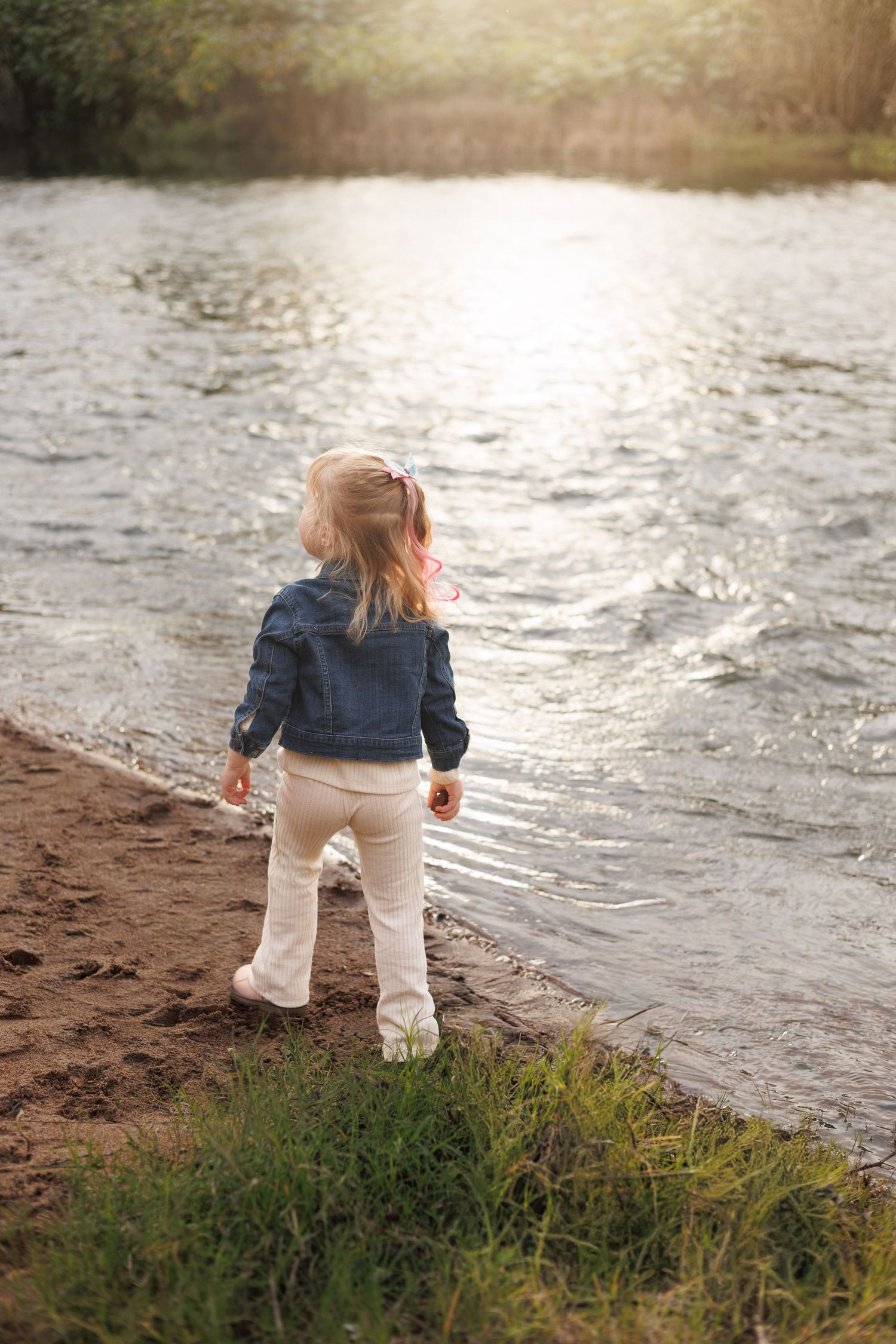 Toddler girl in denim jacket standing on sandy riverbank gazing at sunlit shimmering water.