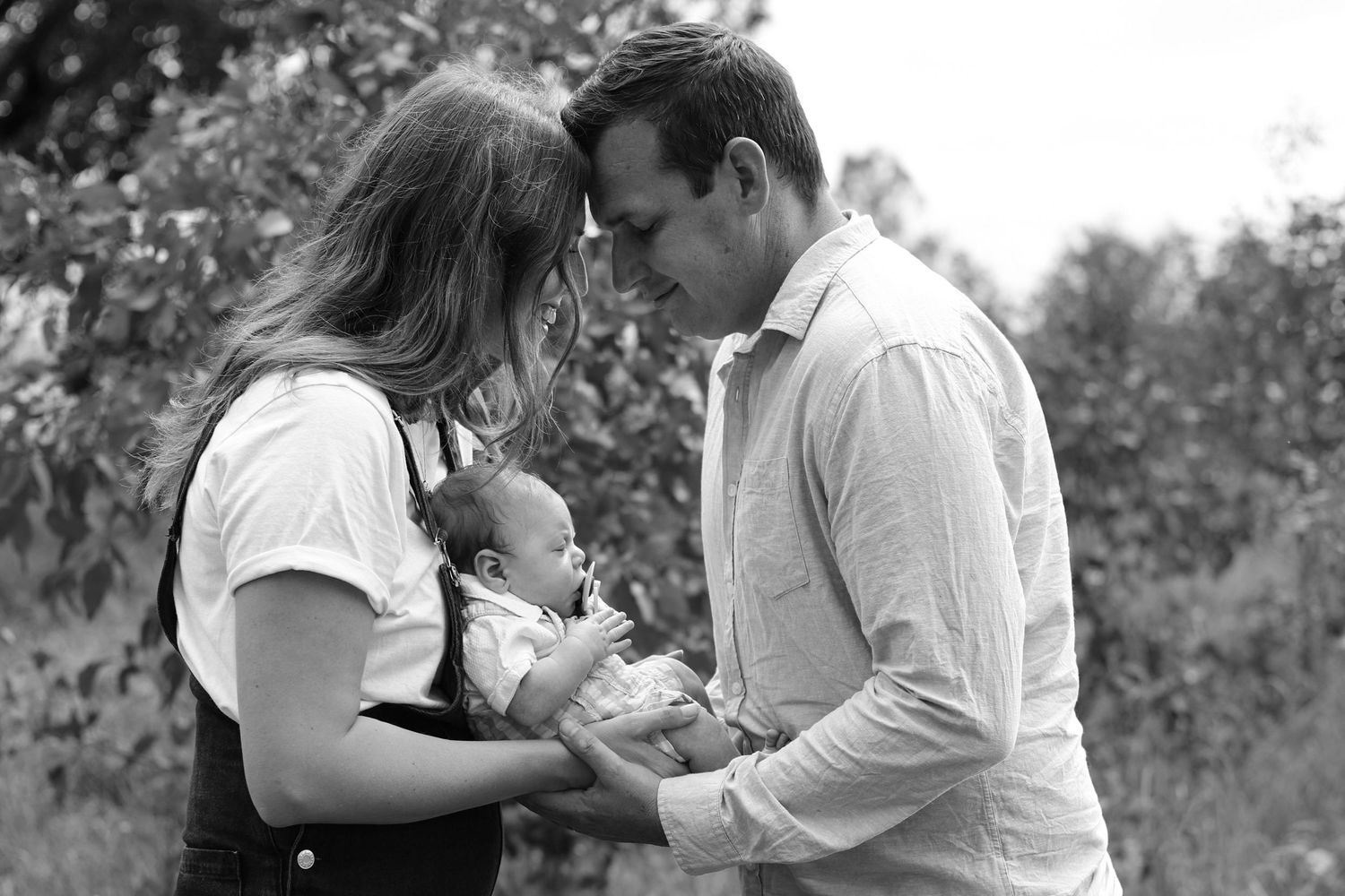 Black and white photo of a couple tenderly looking at their newborn baby outdoors among trees.