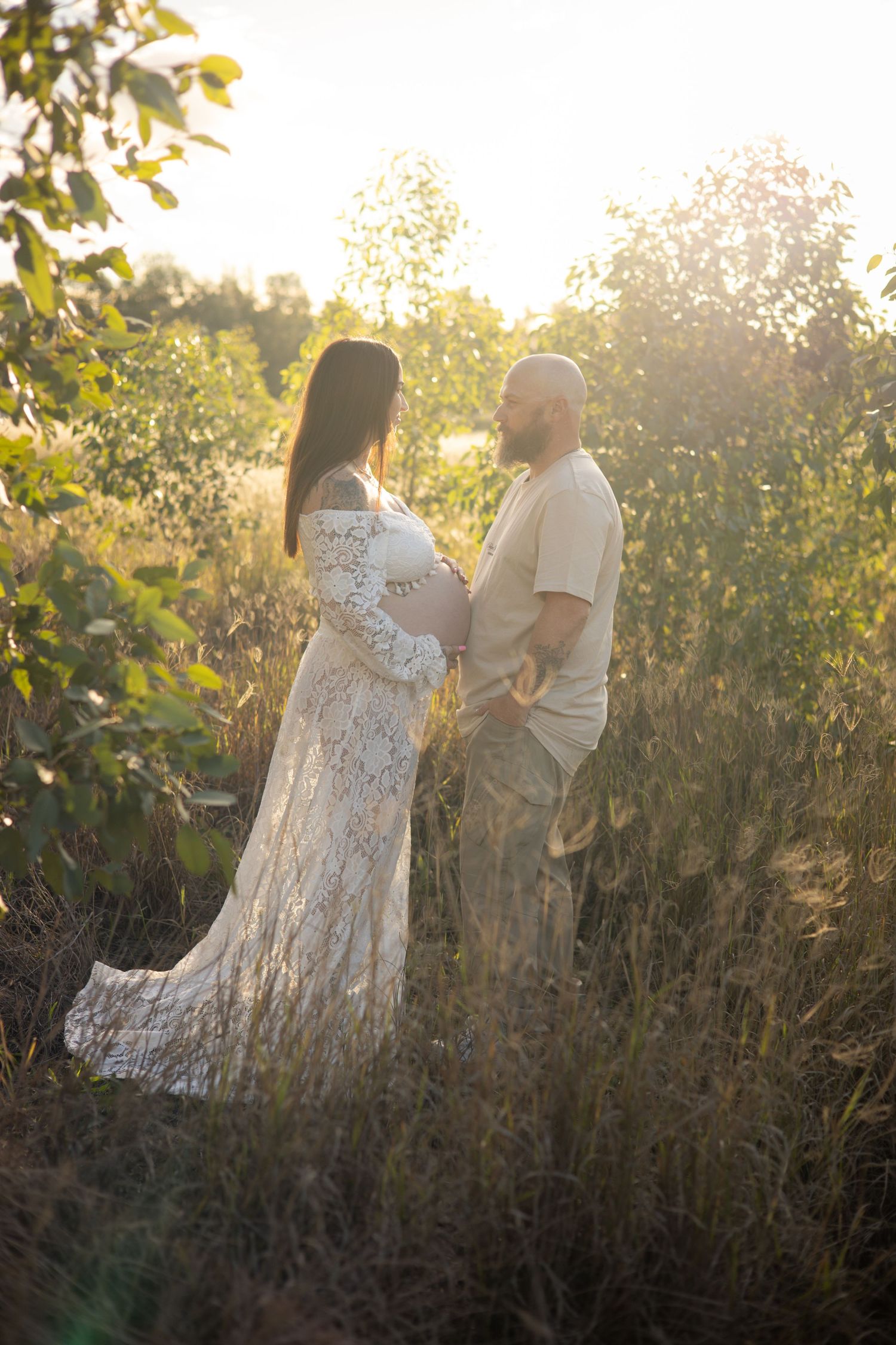 Pregnant couple standing in golden sunlit field, woman in white lace gown cradling baby bump, man in beige outfit facing her.