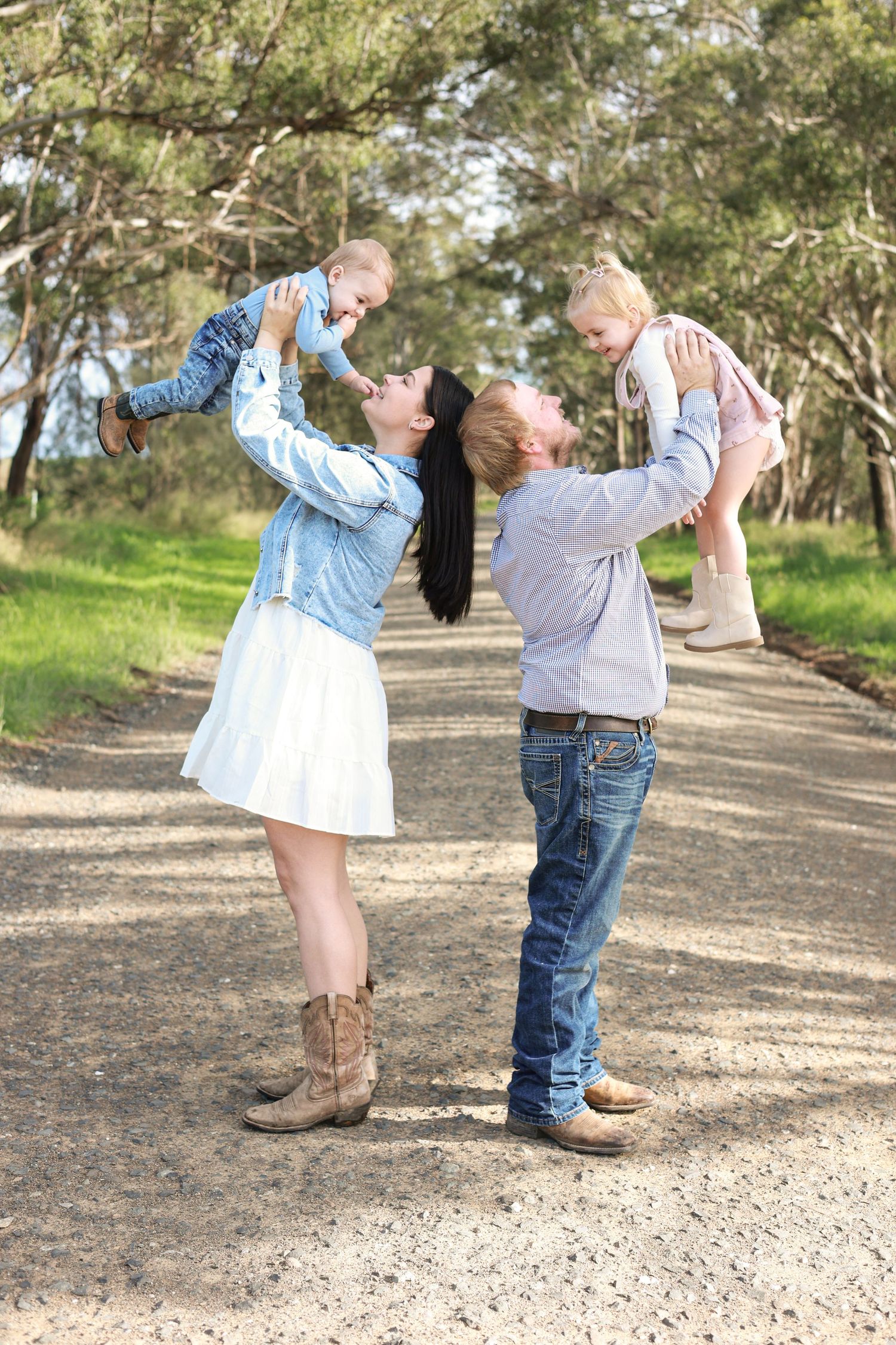 Happy family with two young children playing outdoors on a dirt path surrounded by green trees.