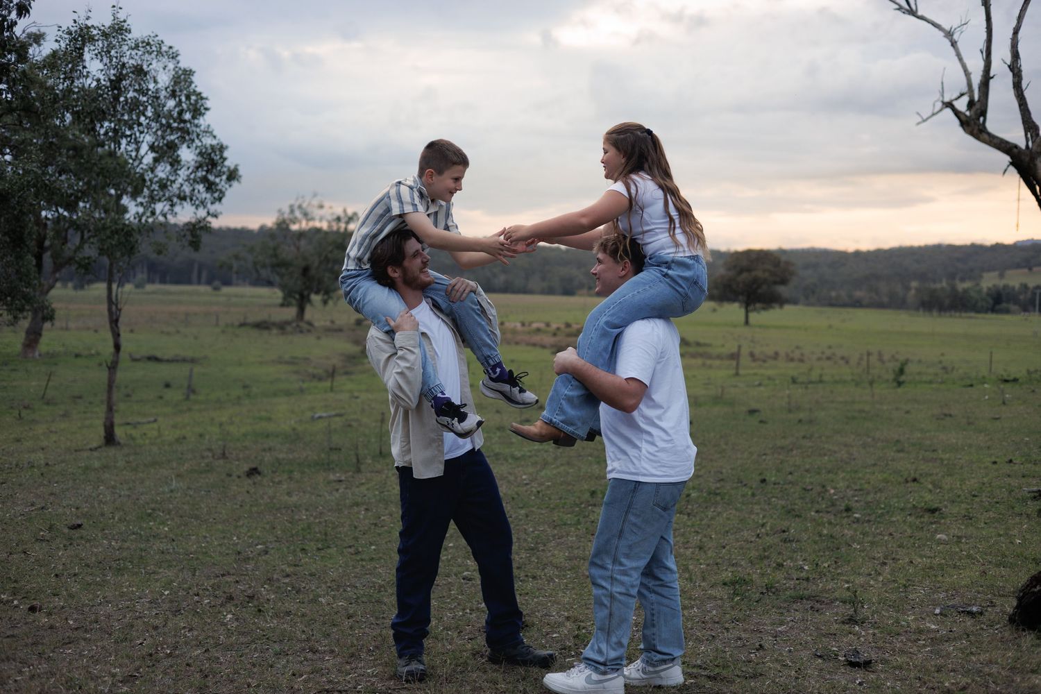 Two adults carry children on their shoulders in a green field at dusk, holding hands in a joyful family moment.
