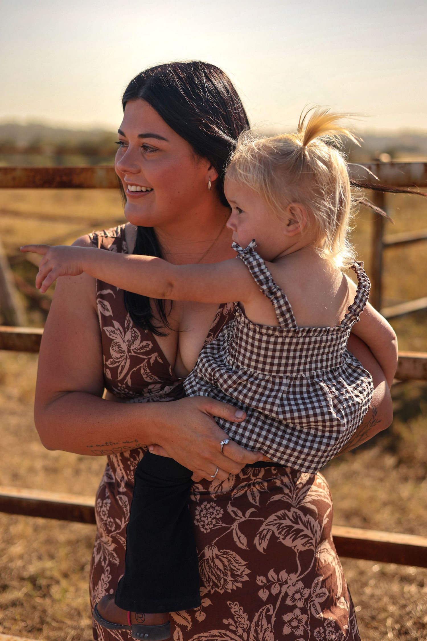 A smiling woman in a floral dress holds a blonde toddler girl outdoors near a rustic fence at golden hour.