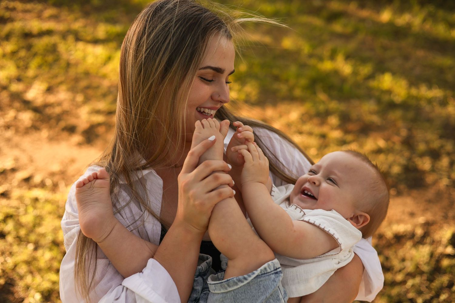 A young mother laughs joyfully while holding her happy baby outdoors in warm golden sunlight.