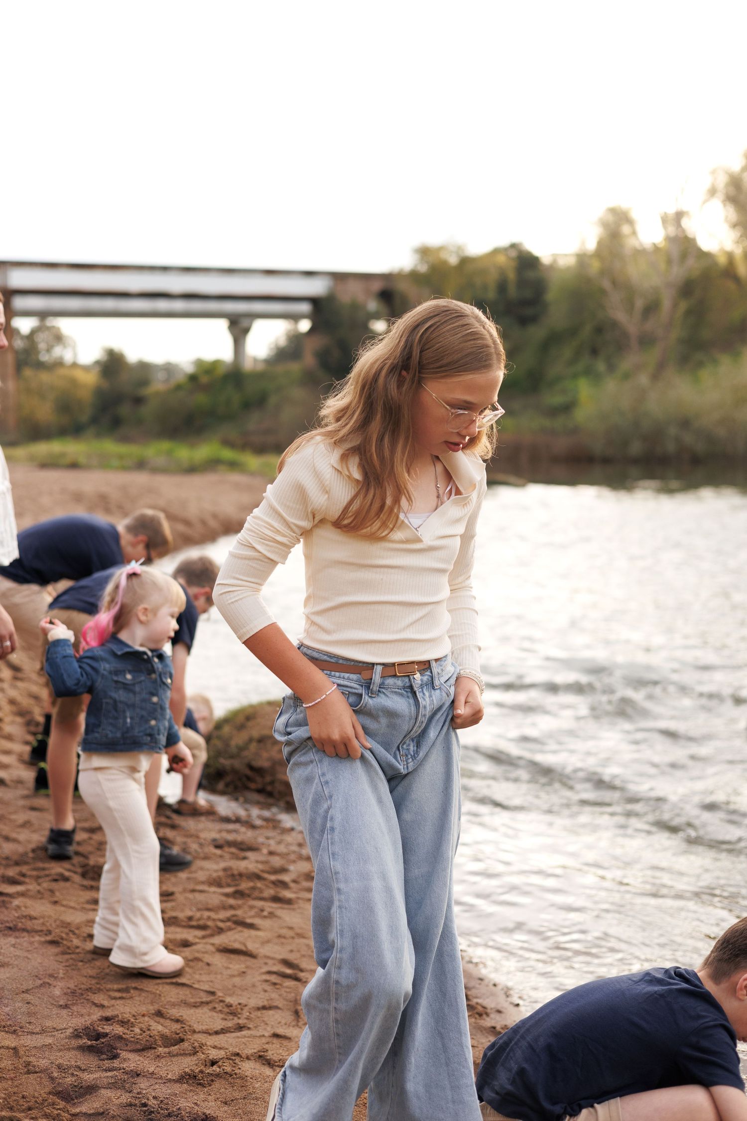 A young girl in wide-leg jeans and glasses stands on a sandy riverbank with children playing behind her.