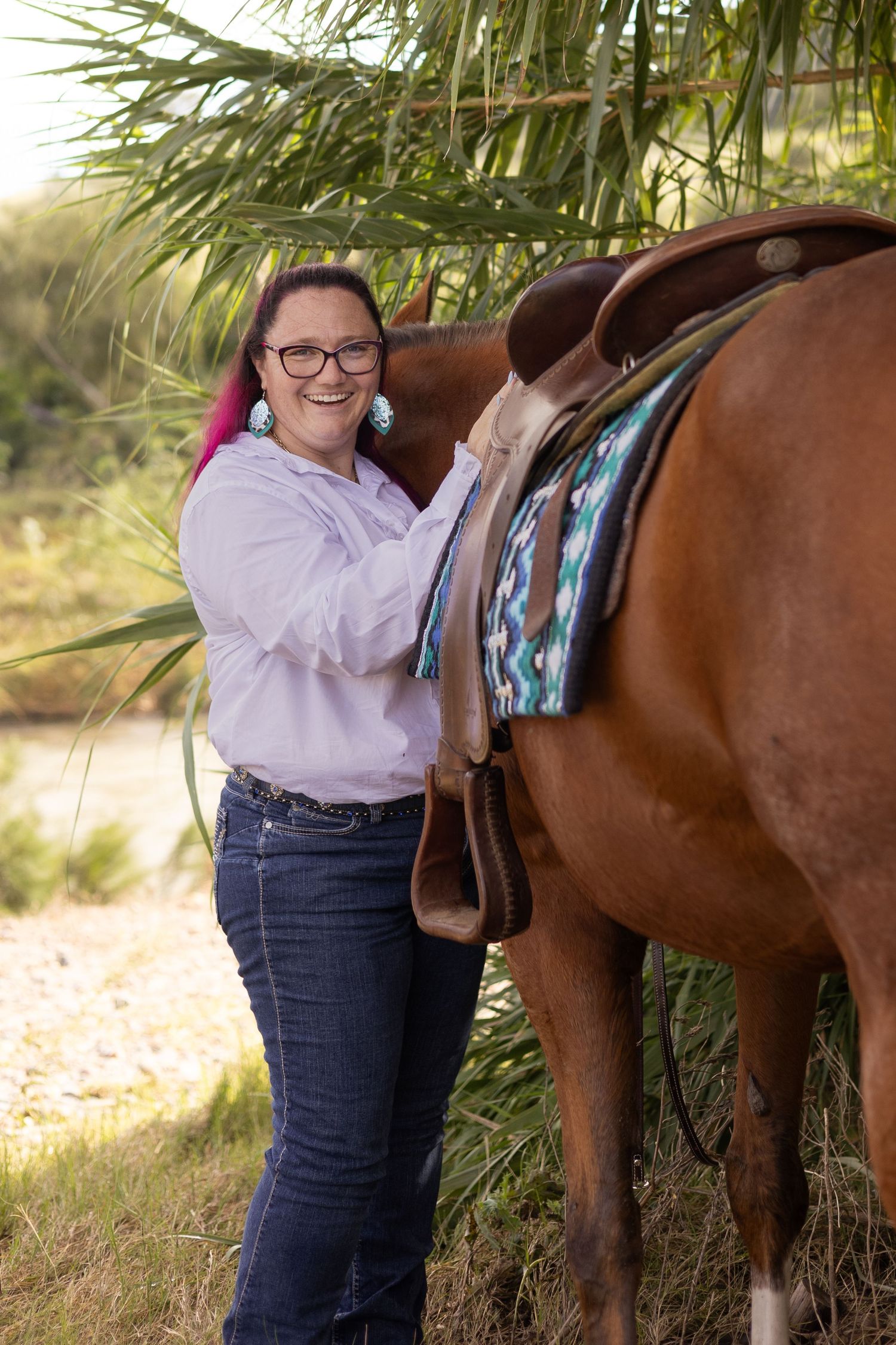 A person in a light shirt and jeans adjusts a saddle on a brown horse under palm trees in an outdoor setting.