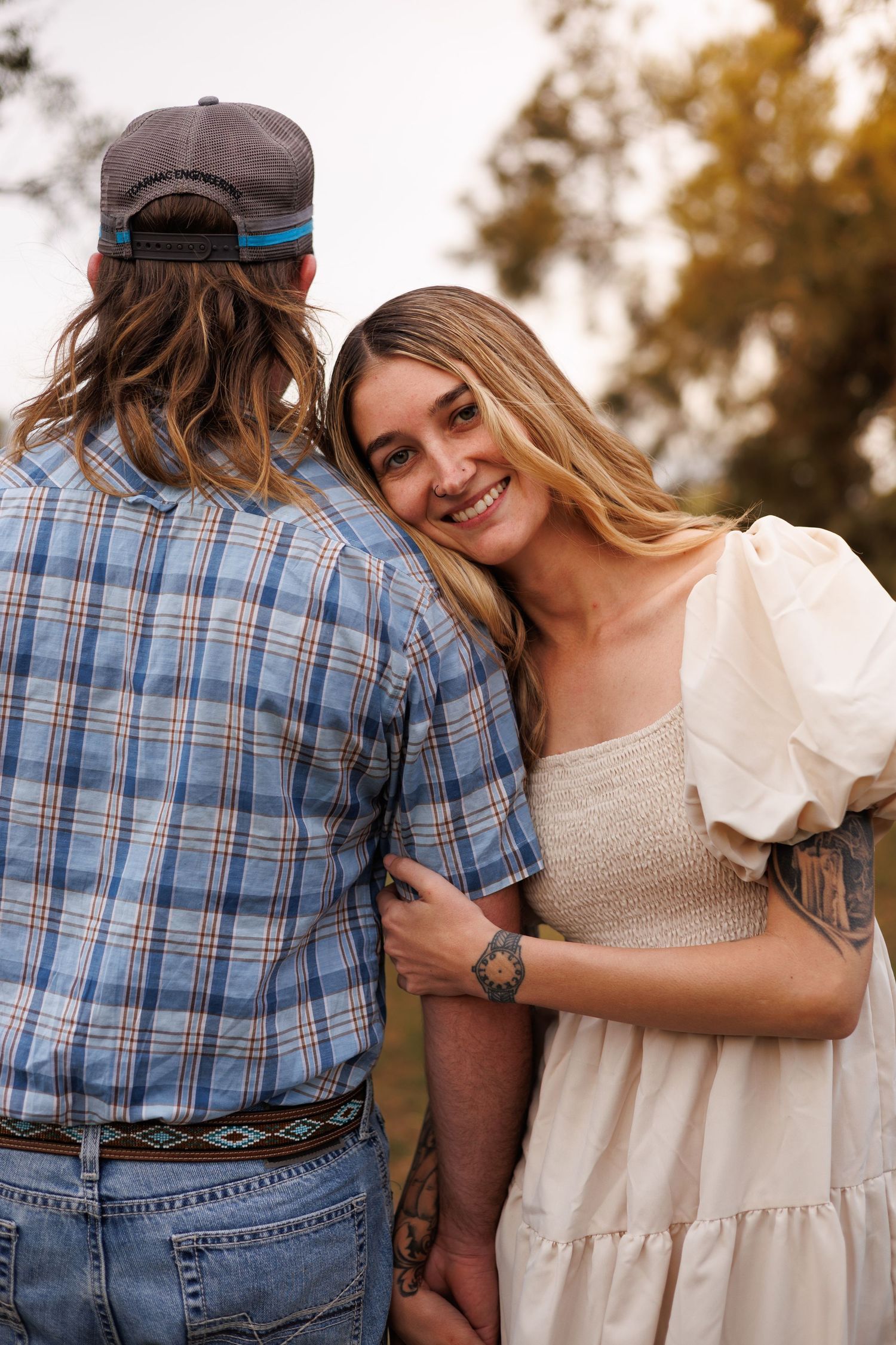 A smiling woman in a white dress embraces a man in a plaid shirt outdoors, holding hands in a romantic couple portrait.
