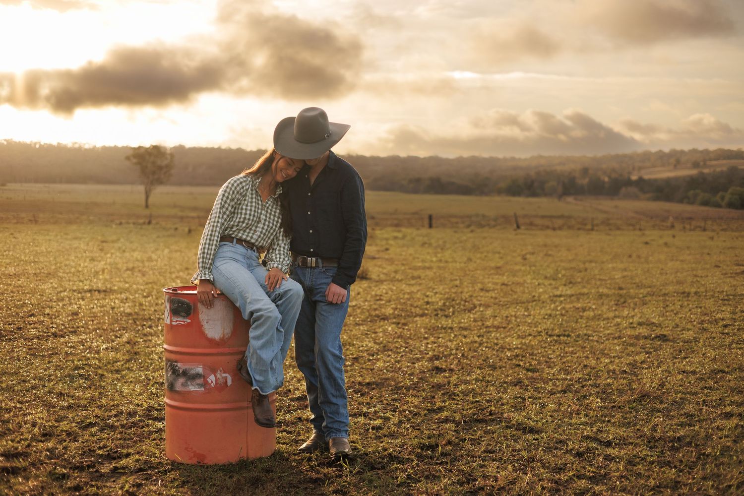 Couple in cowboy hats sharing a romantic moment leaning on a red barrel in a golden sunset pasture.