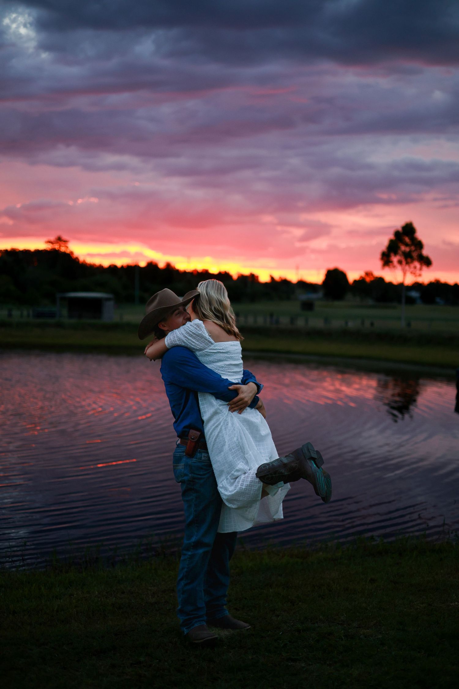 Silhouetted couple embraces by a lake at sunset with dramatic purple and orange sky reflecting on the water.