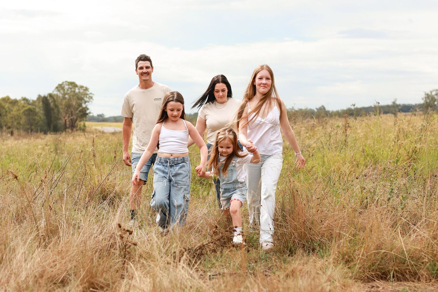 Happy family of five walking through a golden field on a cloudy day, parents and three daughters holding hands.