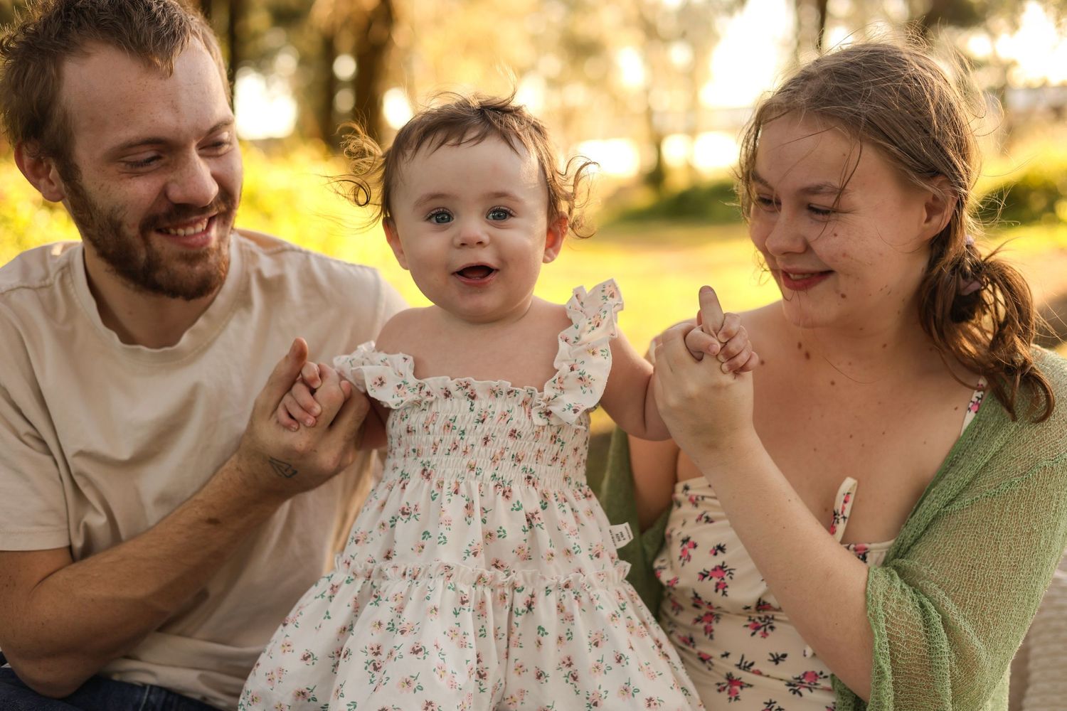 Happy family with smiling baby girl in floral dress held by parents outdoors in sunny park setting.