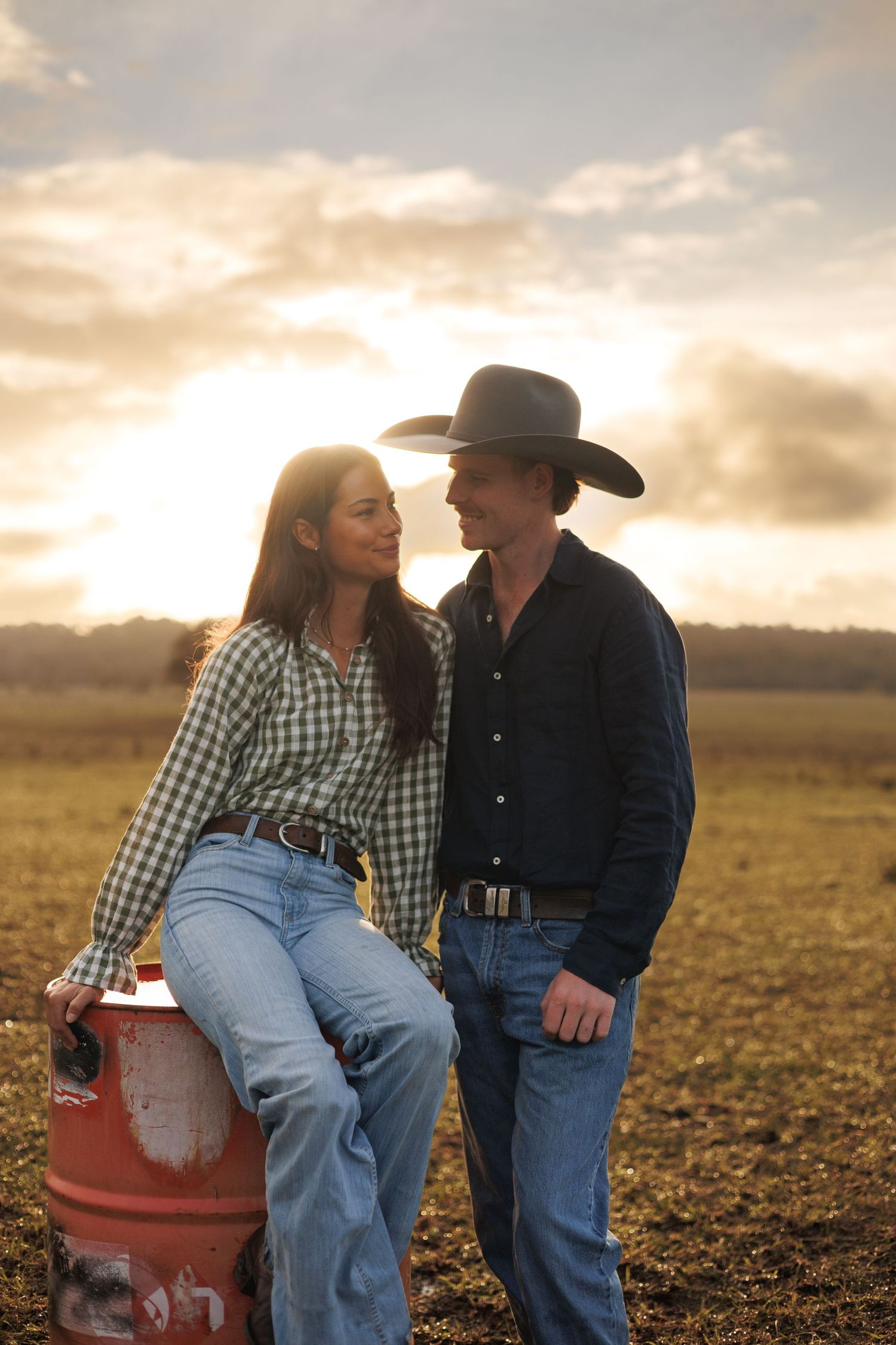 A couple in western wear shares a romantic moment at sunset in a farm field, woman leaning on a red barrel.