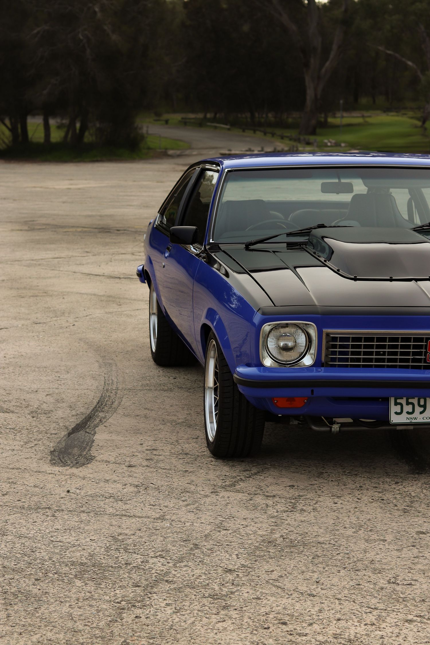 A classic blue Holden Torana LX hatchback with chrome wheels parked on a dirt path surrounded by trees.