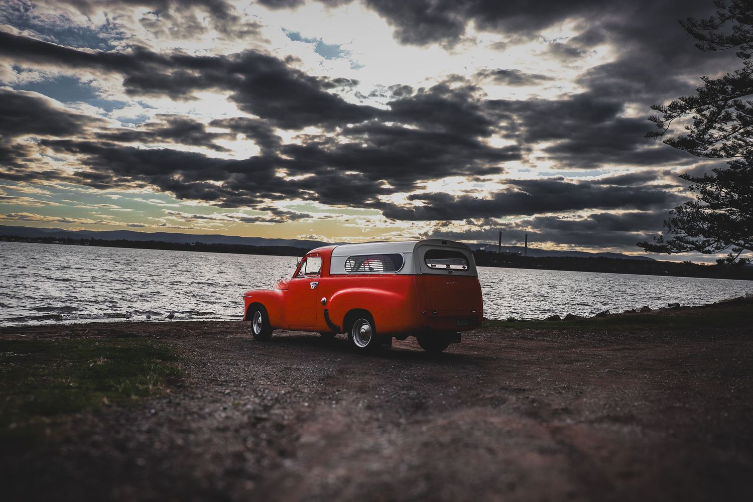 A bright orange vintage pickup truck silhouetted against a dramatic cloudy sunset sky over the ocean horizon.