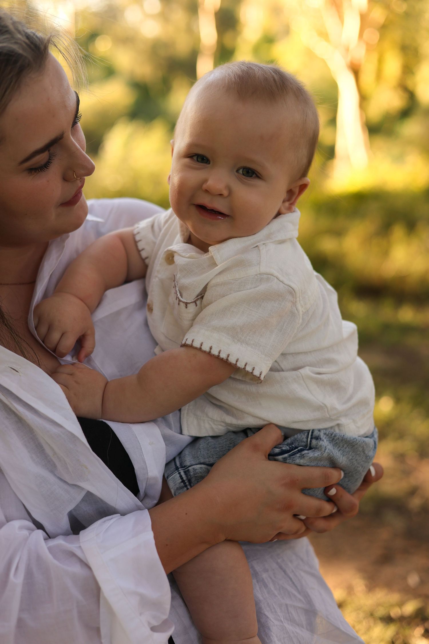 A smiling baby in a white shirt and jeans being held by a woman in a white dress outdoors in a sunny park.