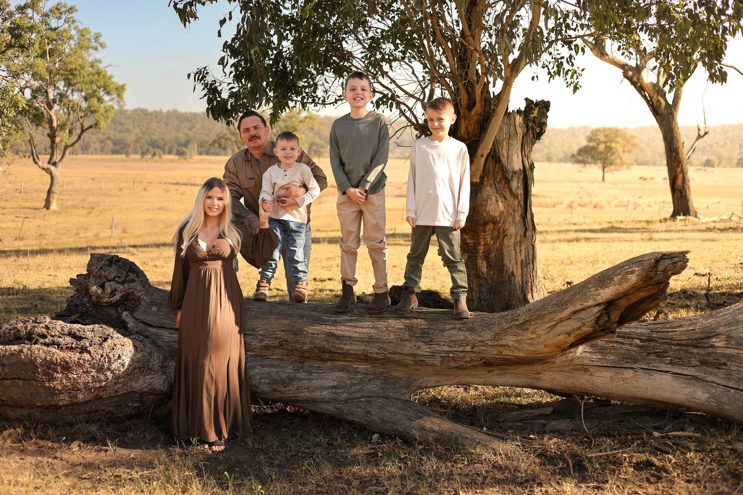 Family of five posing near a fallen log in a golden Australian outback field with gum trees at sunset.