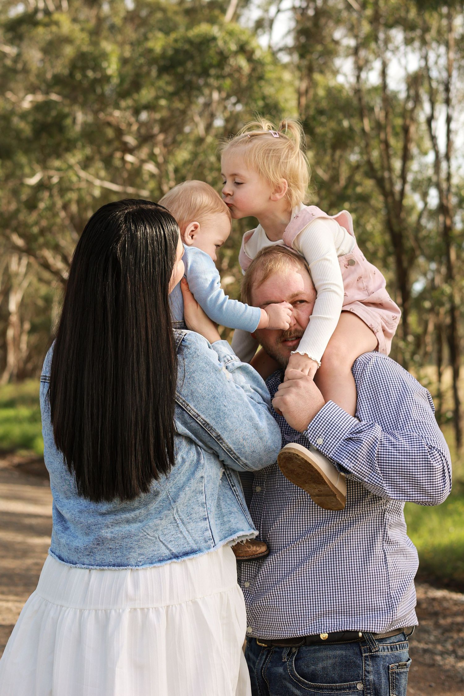Family of four enjoying outdoor time, parents holding baby and toddler girl in a tree-lined park path.
