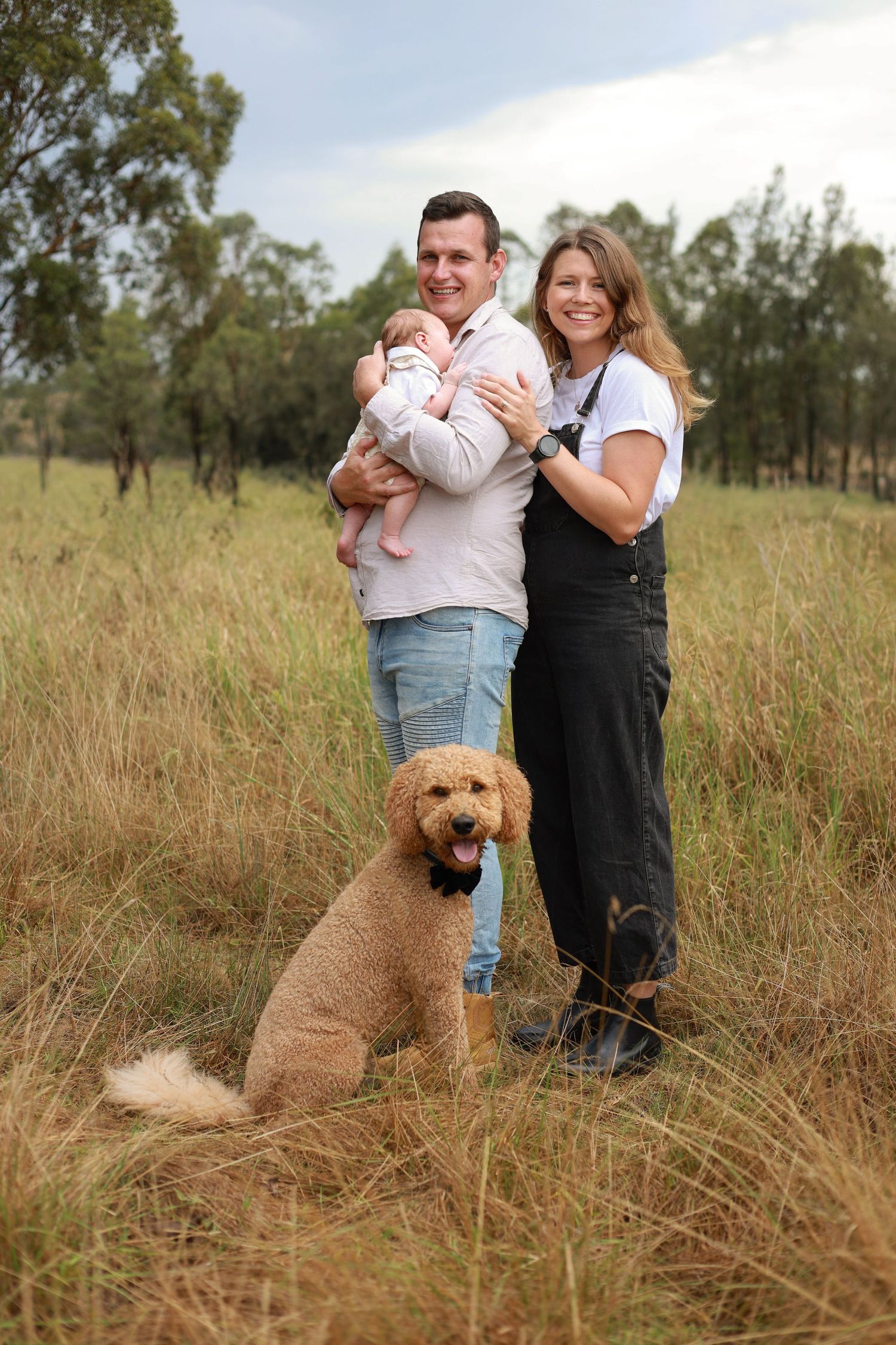 Young family with baby and golden doodle dog posing in a grassy field with eucalyptus trees in rural Australia.