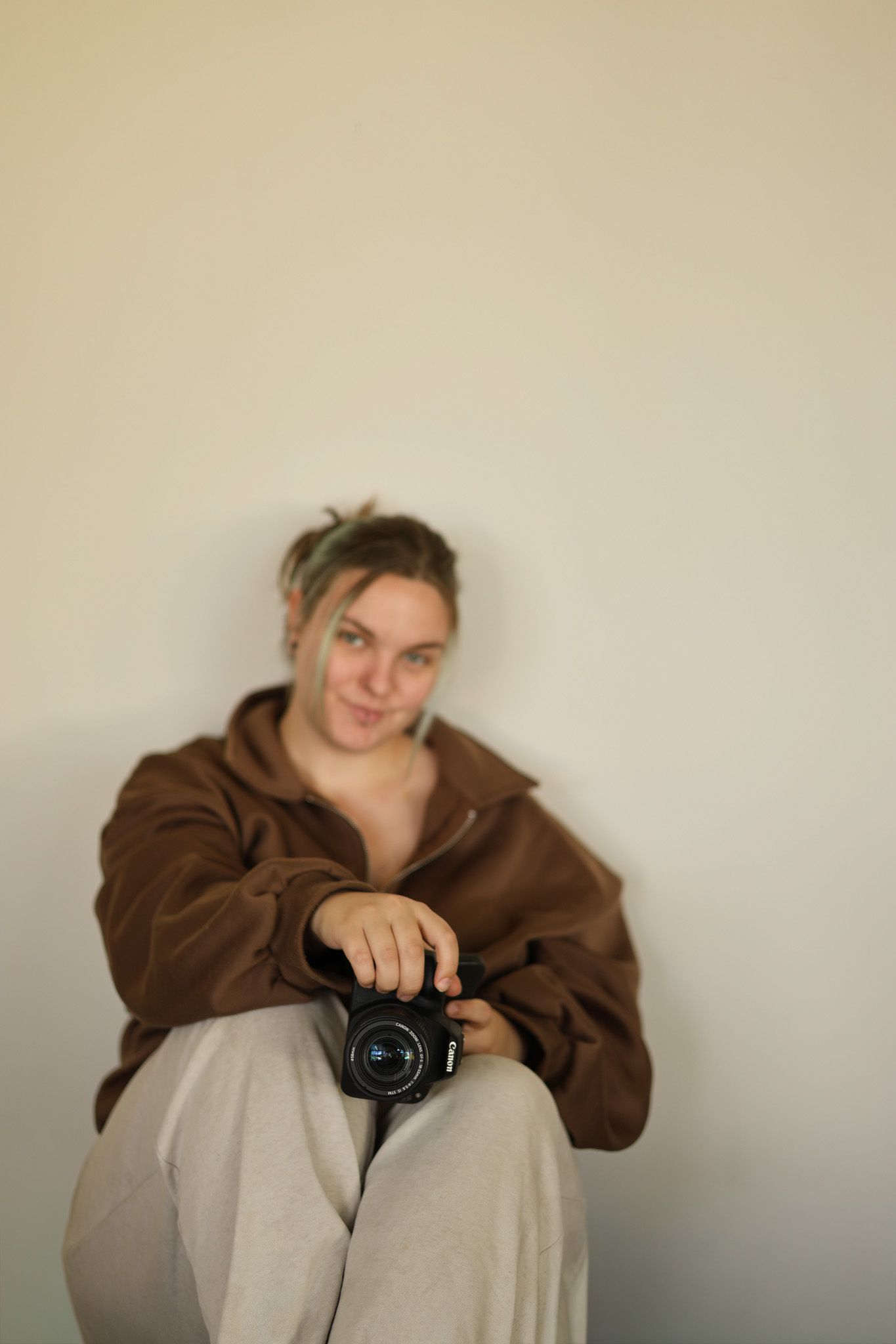 Young woman in brown jacket sitting against wall holding a black camera, smiling softly at viewer.