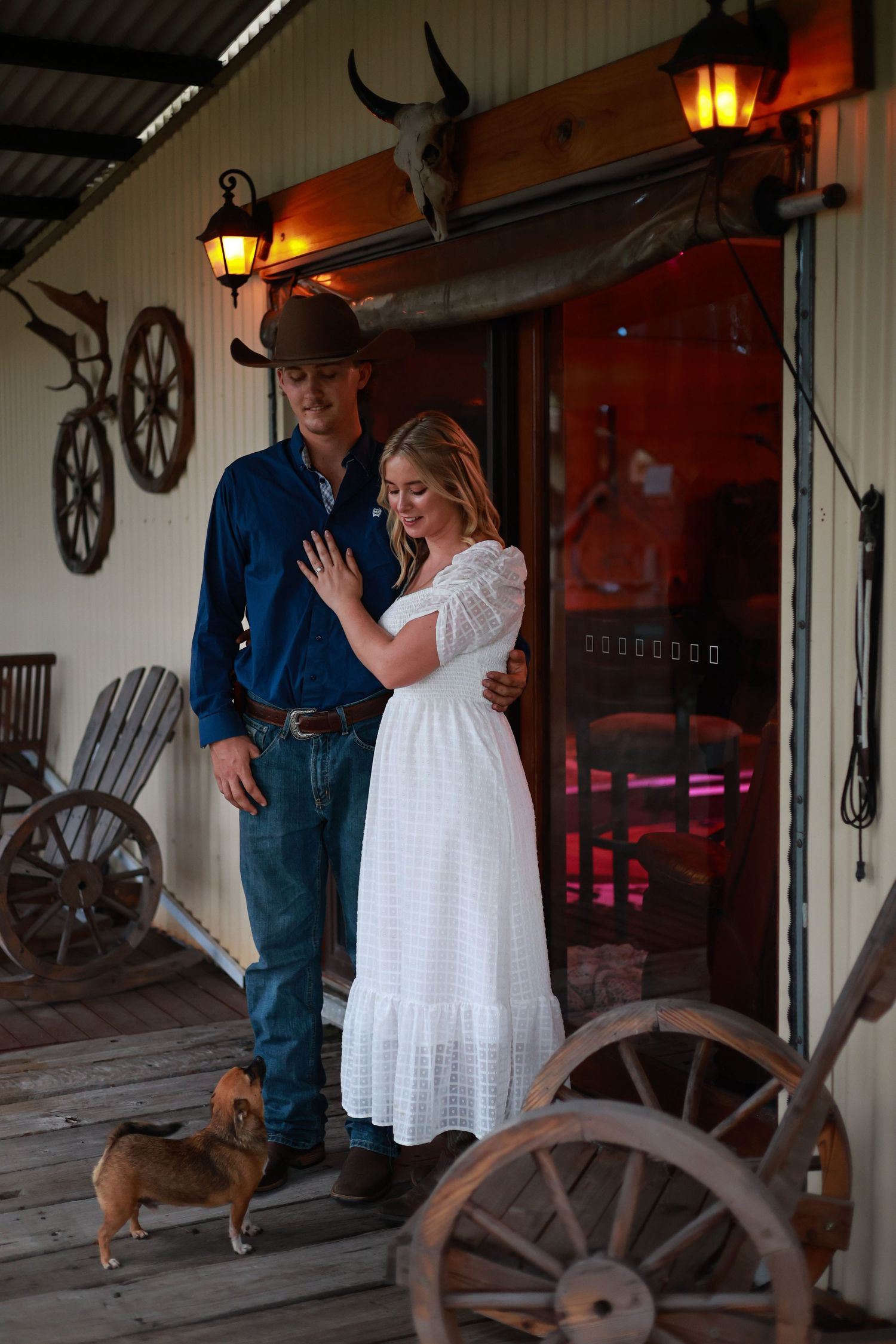 A rustic western-themed porch scene with a couple in cowboy attire and a small dog, decorated with wagon wheels and lanterns.