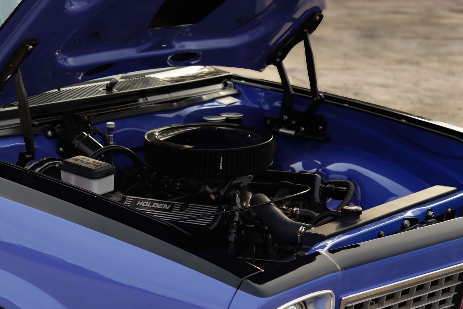 Close-up view of a dark blue classic car hood with chrome trim and intake details being mechanically serviced.