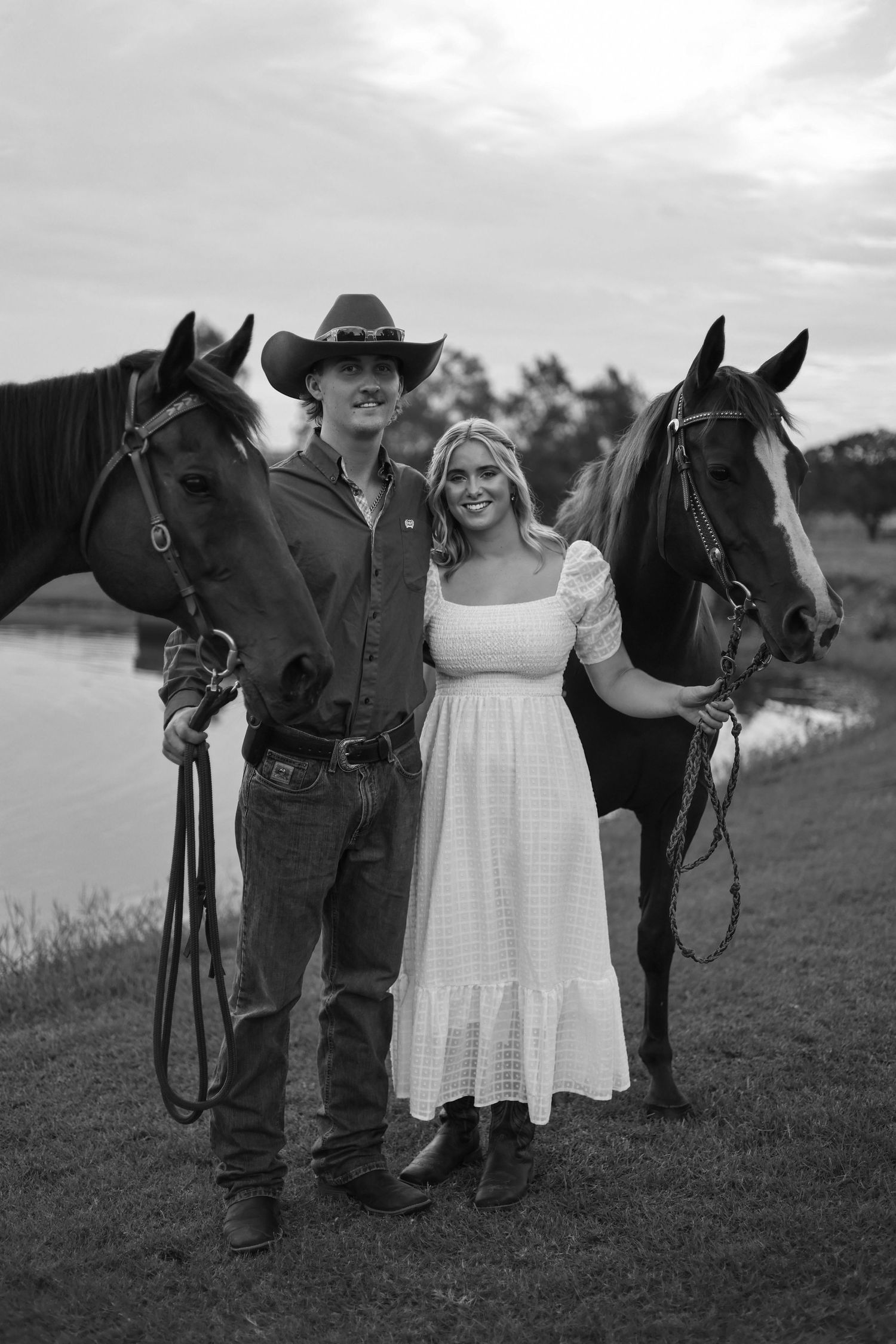 A black and white engagement photo shows a couple in country attire standing with their horses in a rustic field.