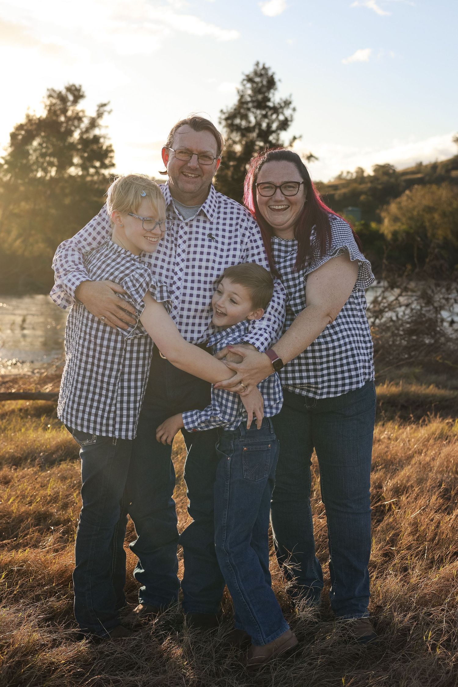 A family of four wearing coordinated blue and white checkered shirts poses together in a golden field at sunset.