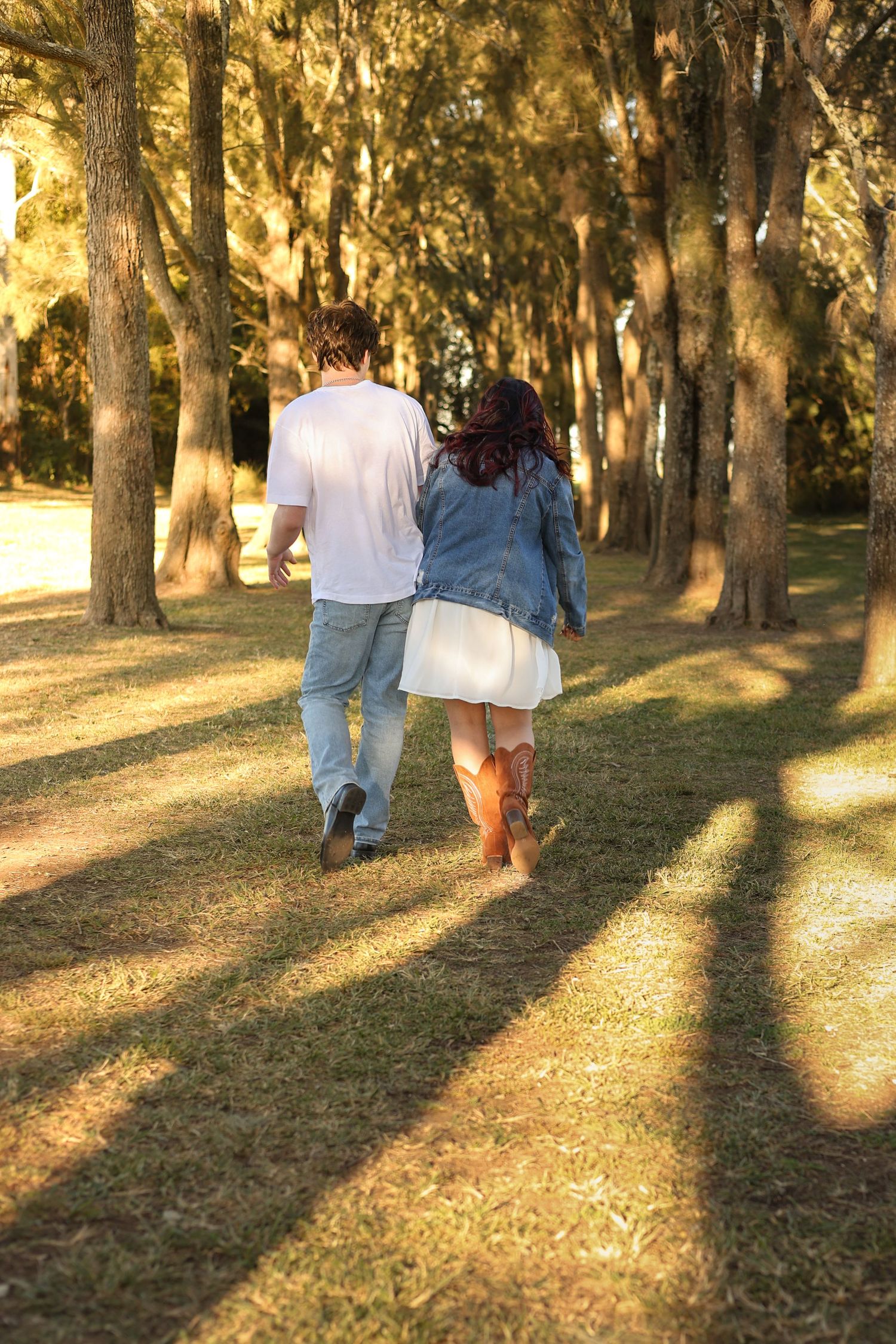 Couple walking hand in hand through a sunlit tree-lined park path, woman in white dress and denim jacket with cowboy boots.