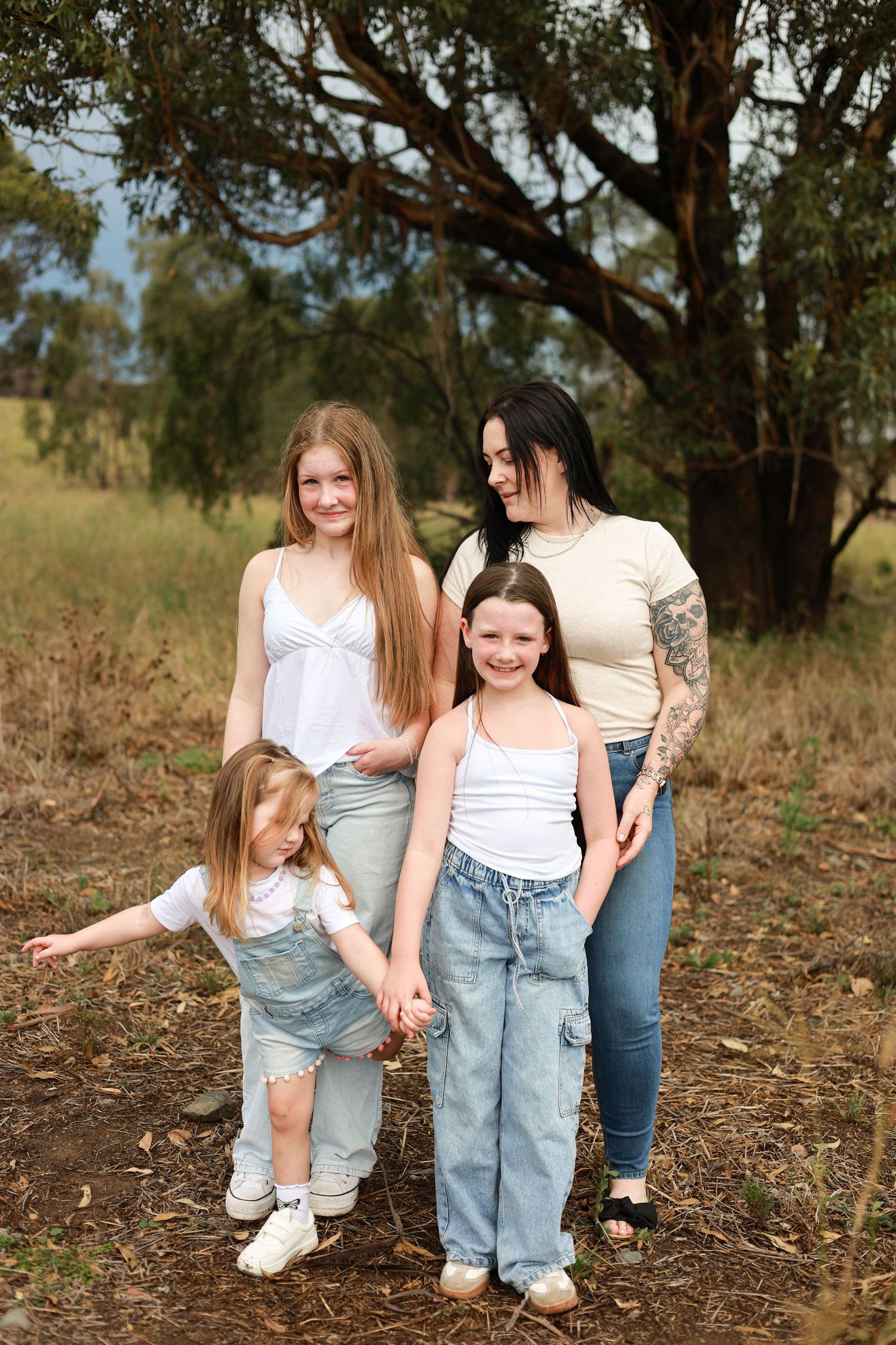 A mother and three daughters pose together outdoors in a field with trees, wearing casual summer outfits.