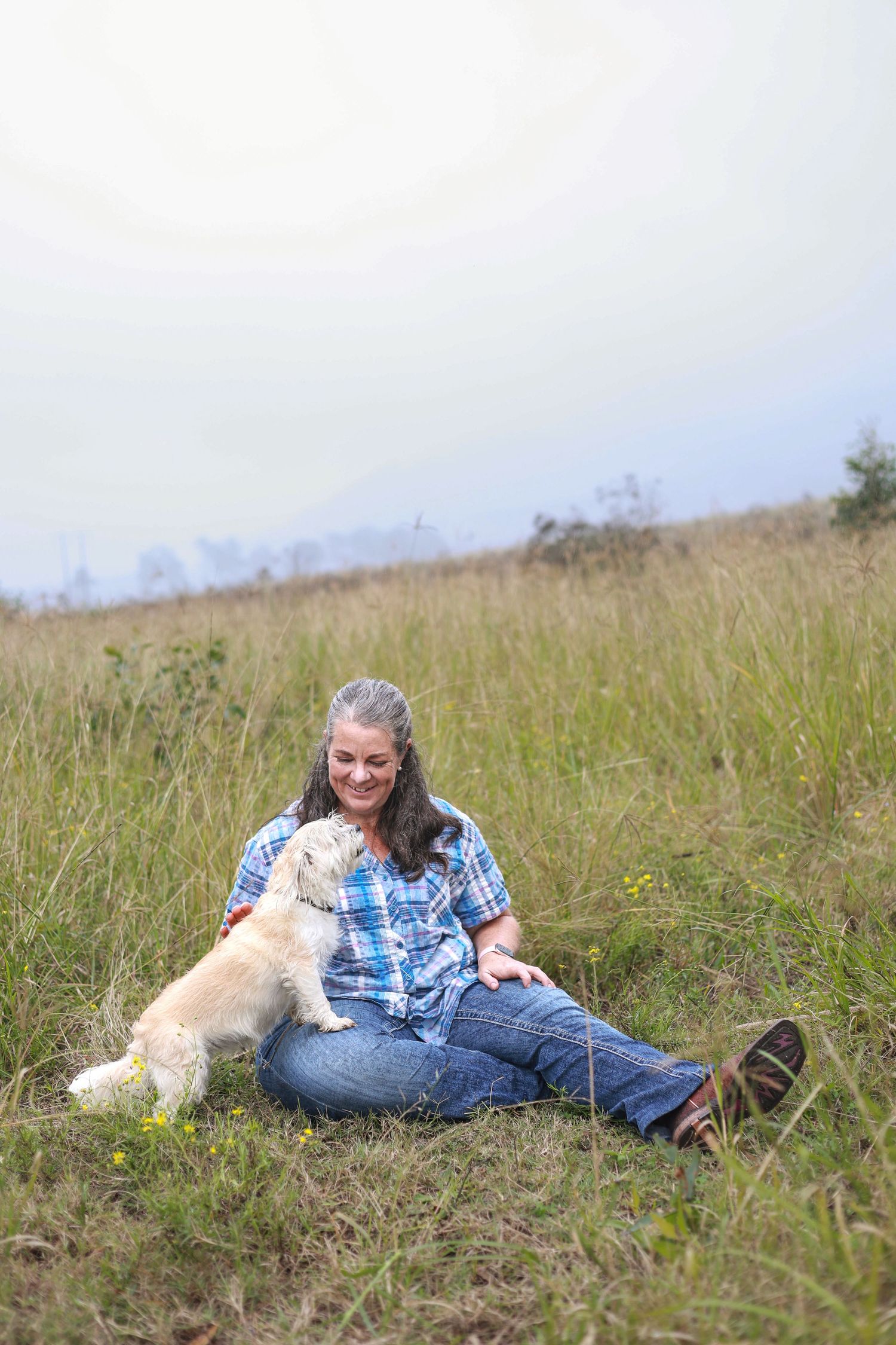Person in blue plaid shirt relaxes in grassy meadow with dog on rainy day.