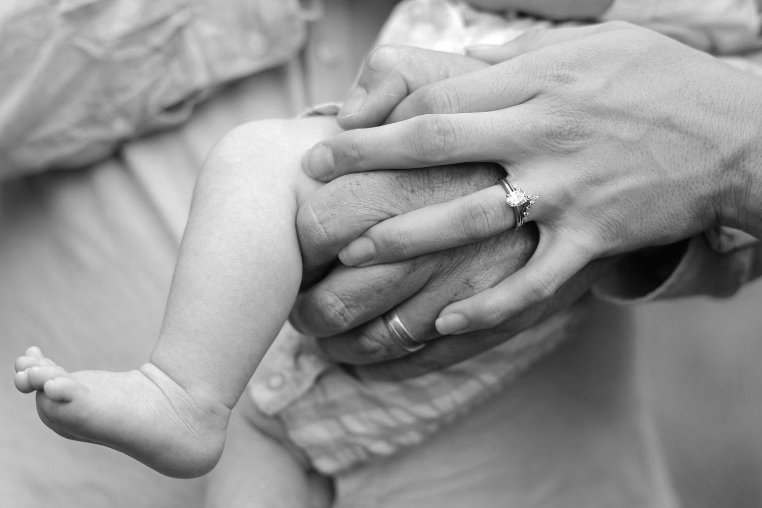 Black and white photo of newborn baby legs cradled in parents hands wearing wedding rings.