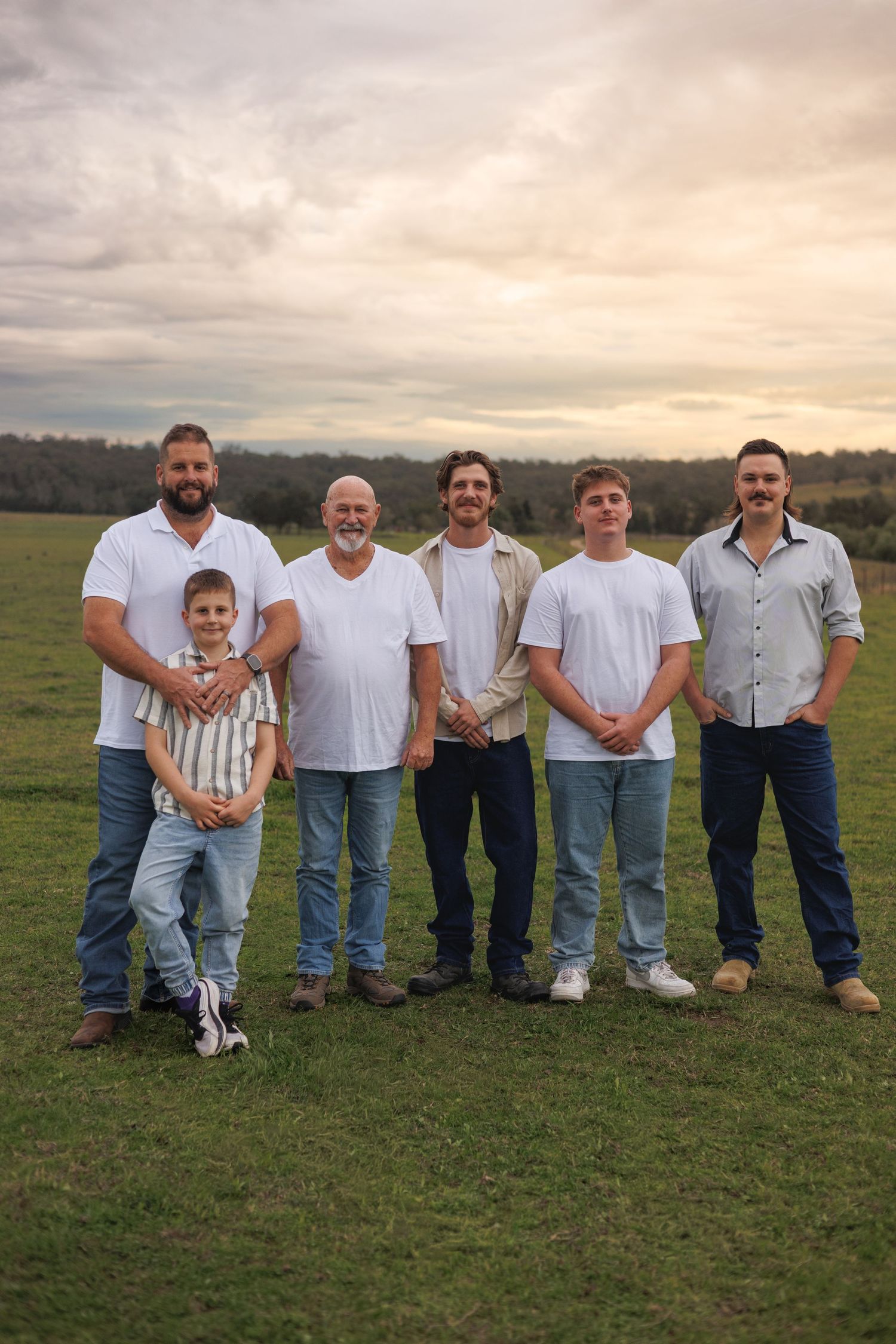Six males of varying ages pose together in a green field under a cloudy sunset sky, wearing white shirts and jeans.