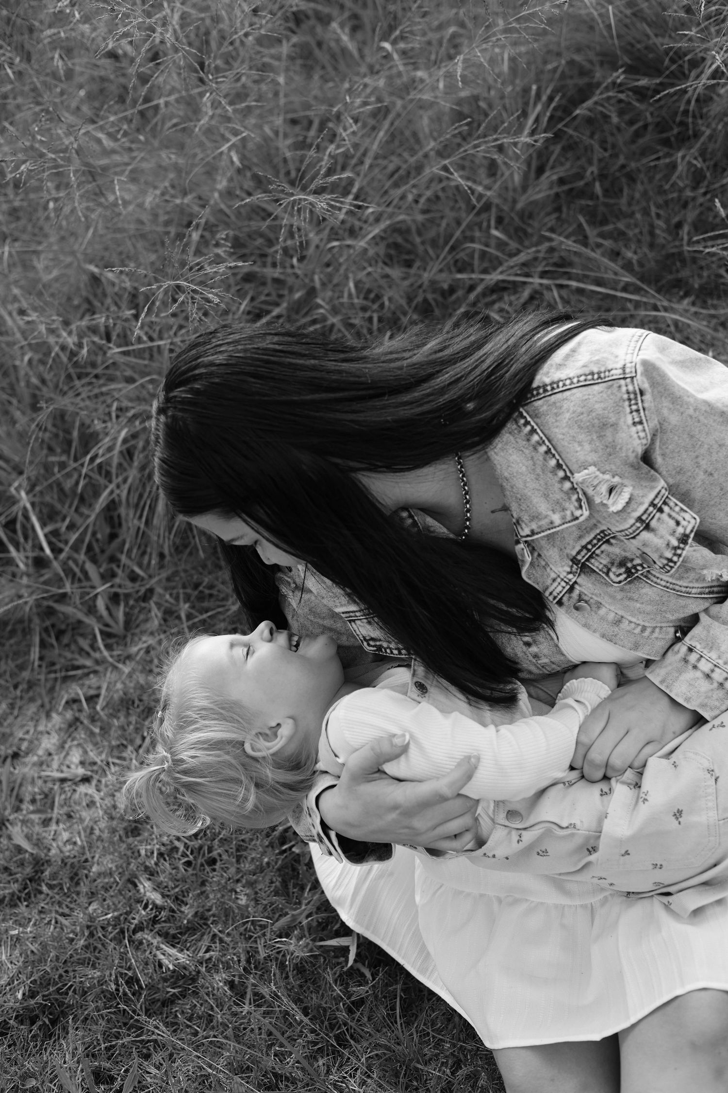 Black and white photo of a dark-haired woman tenderly leaning over a young blonde child in a grassy field.
