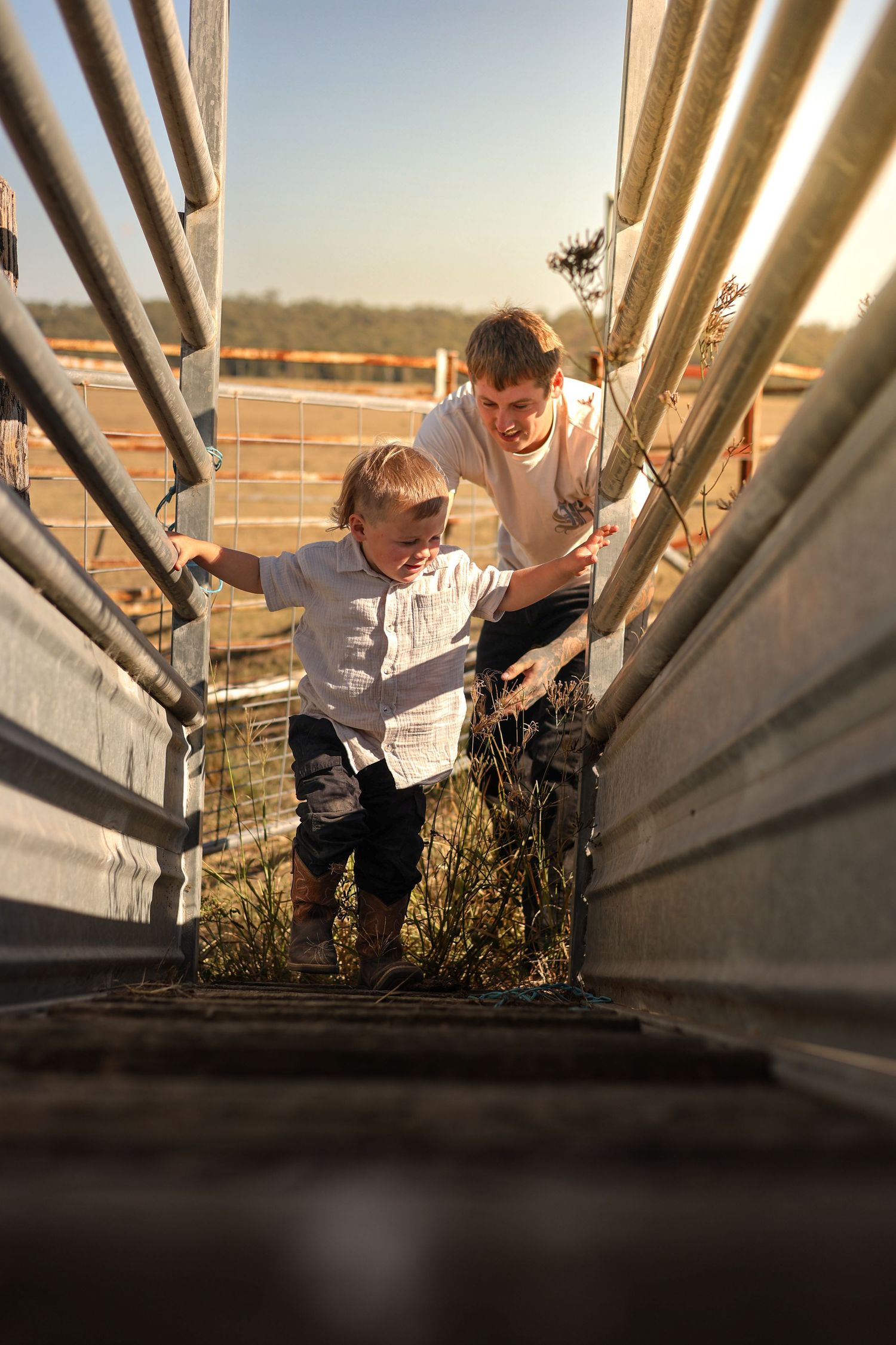 Two boys climbing metal farm yard rails in golden afternoon sunlight, smiling and exploring rural property together.