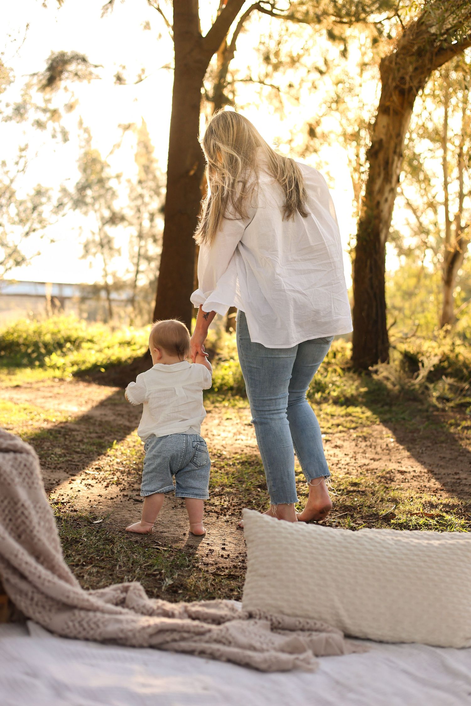A mother helps her toddler walk outdoors in golden sunlight near trees, both wearing casual white tops and jeans.