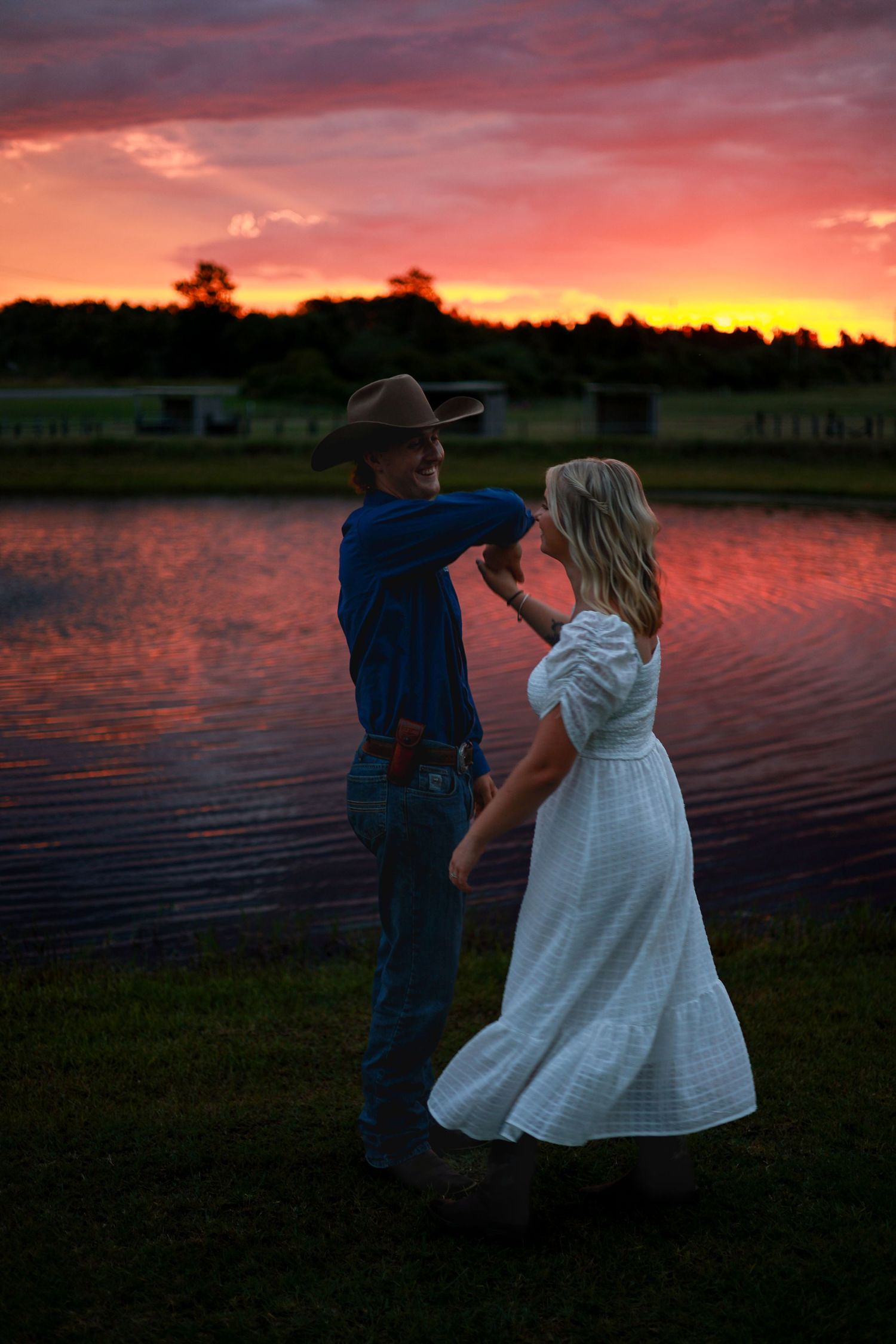 A couple dances together beside a lake at sunset with vibrant orange and pink sky reflecting on the water.