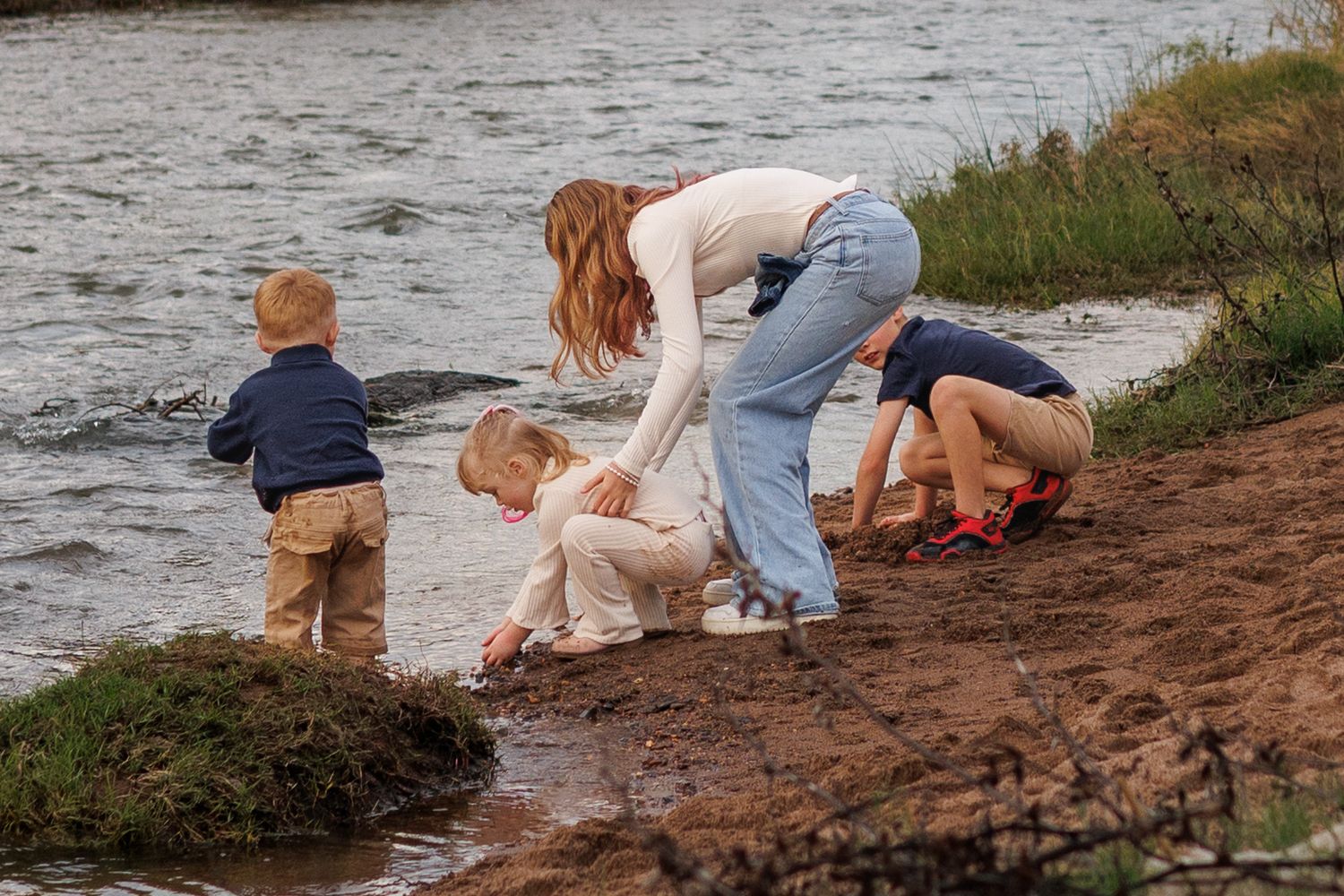A woman helps three young children explore the muddy riverbank edge in a nature outdoor setting.