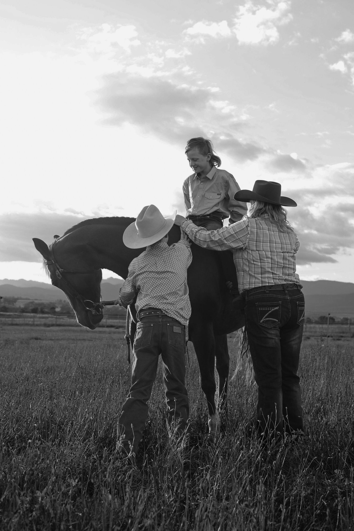 Black and white photo of two cowboys helping a rider mount a dark horse in an open field.