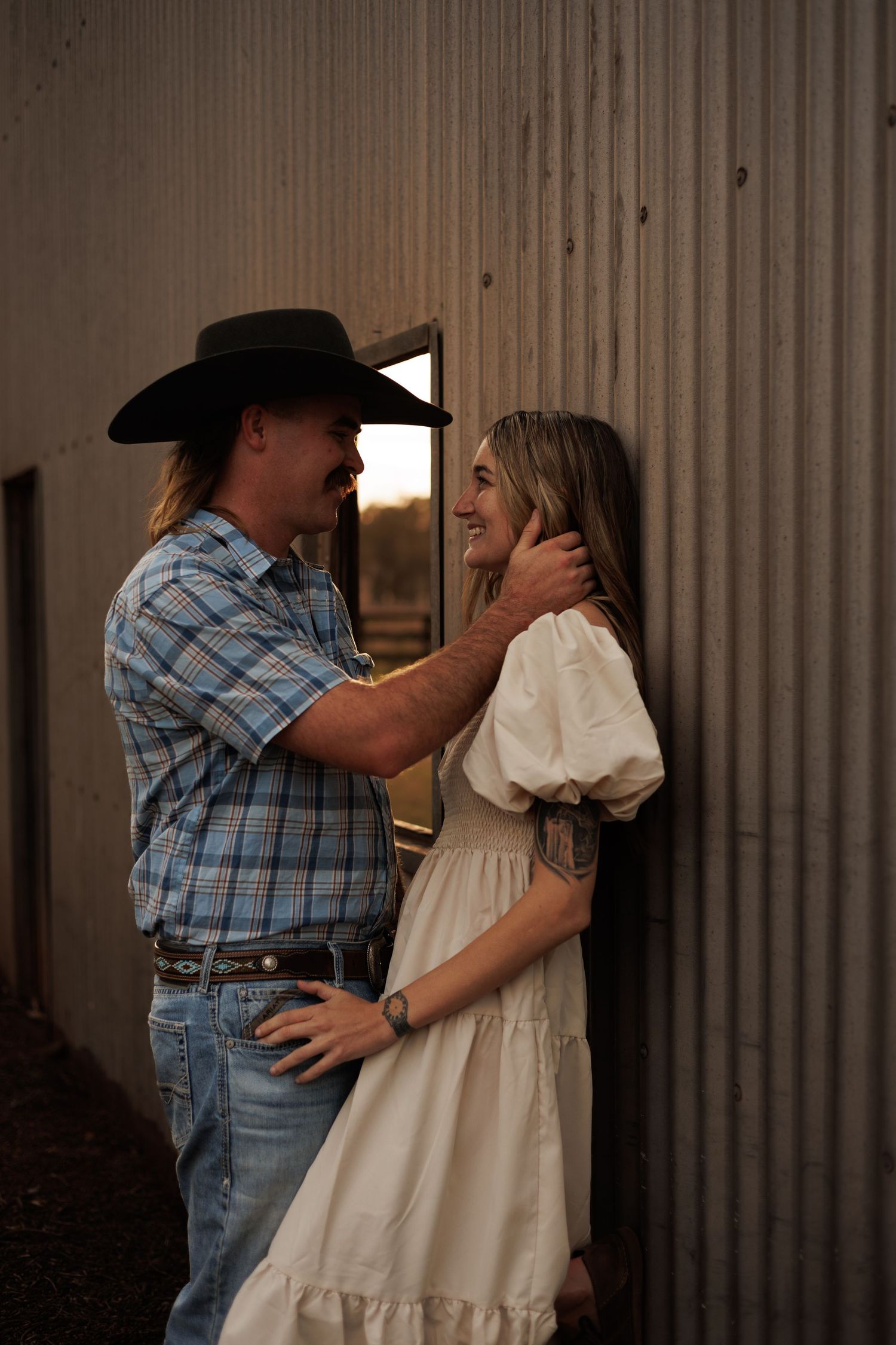 A cowboy in a black hat and plaid shirt tenderly holds a smiling woman in a white dress against a rustic barn wall.