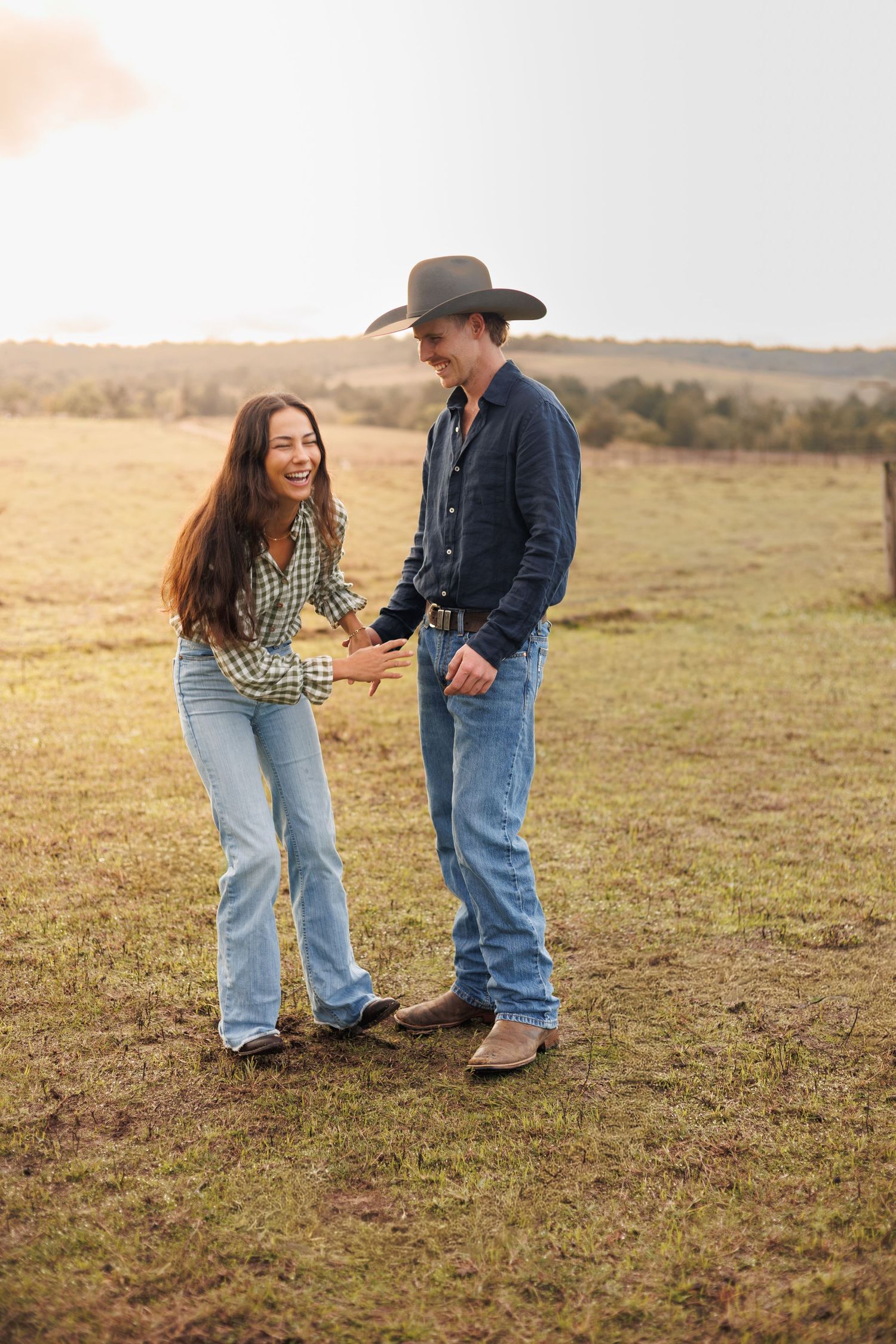 A smiling couple in western attire holding hands in a sunlit green field, wearing jeans and plaid shirts.