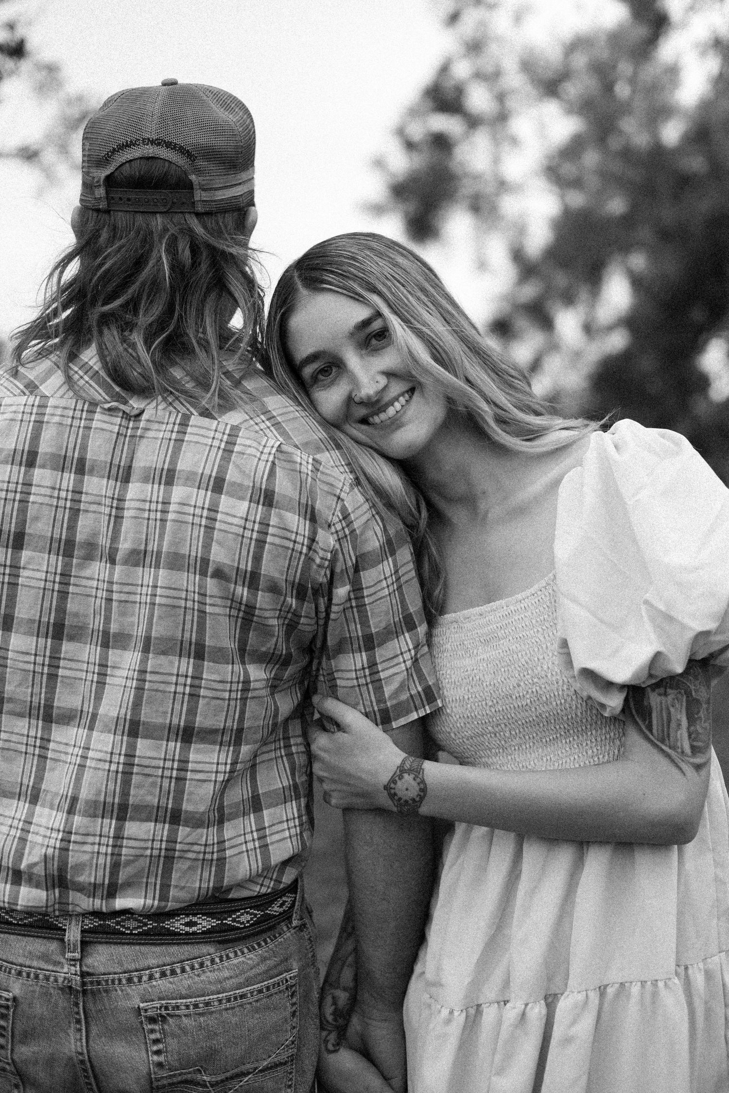 Black and white photo of a smiling woman in white dress leaning on a man in plaid shirt outdoors.