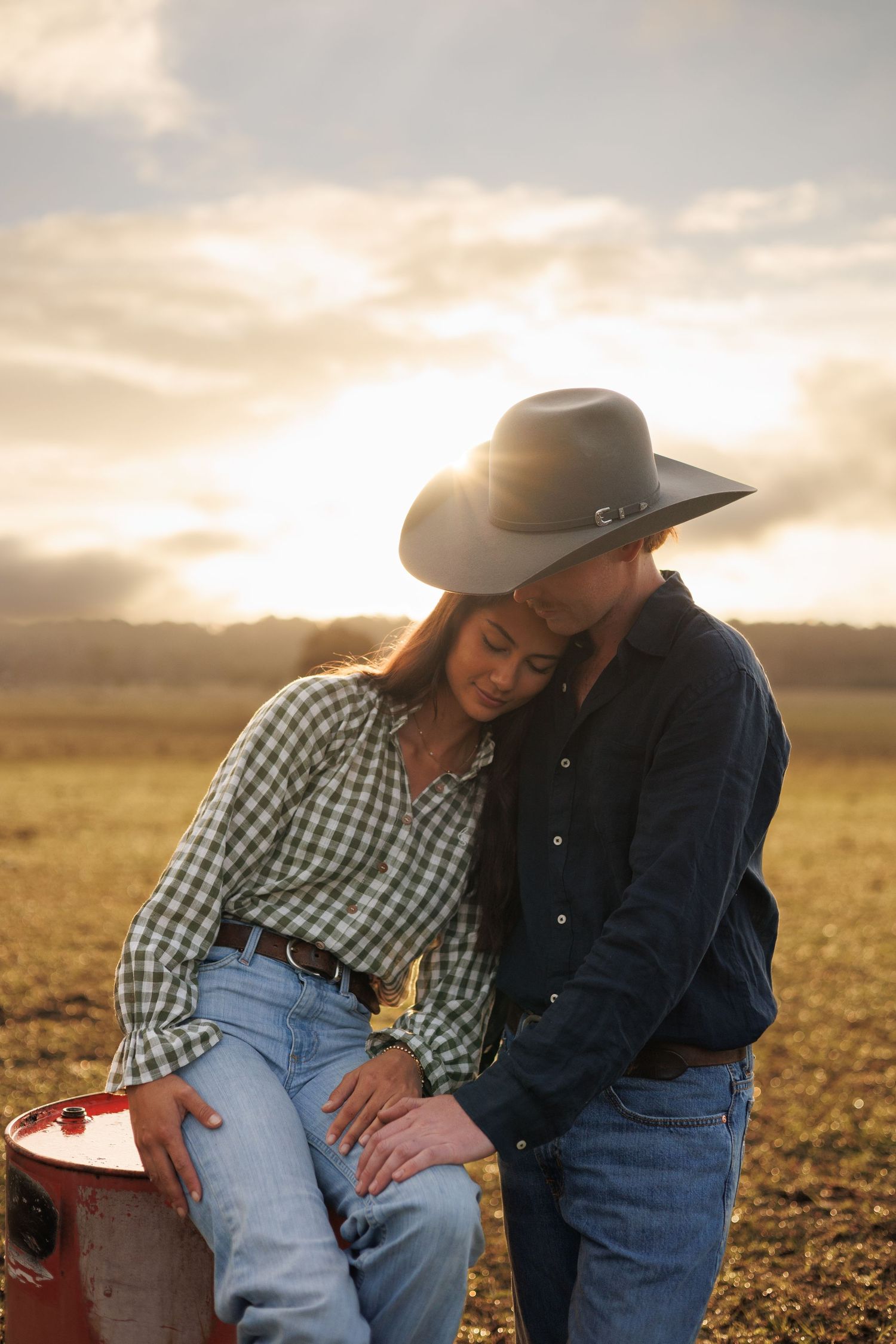 Couple in western wear sharing a tender moment at sunset in an open field, woman in plaid shirt leans on mans shoulder.