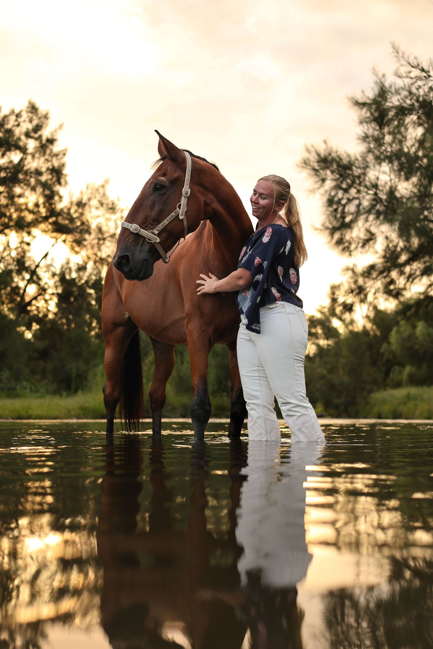 A person in white stands with their brown horse in shallow water during a golden sunset.